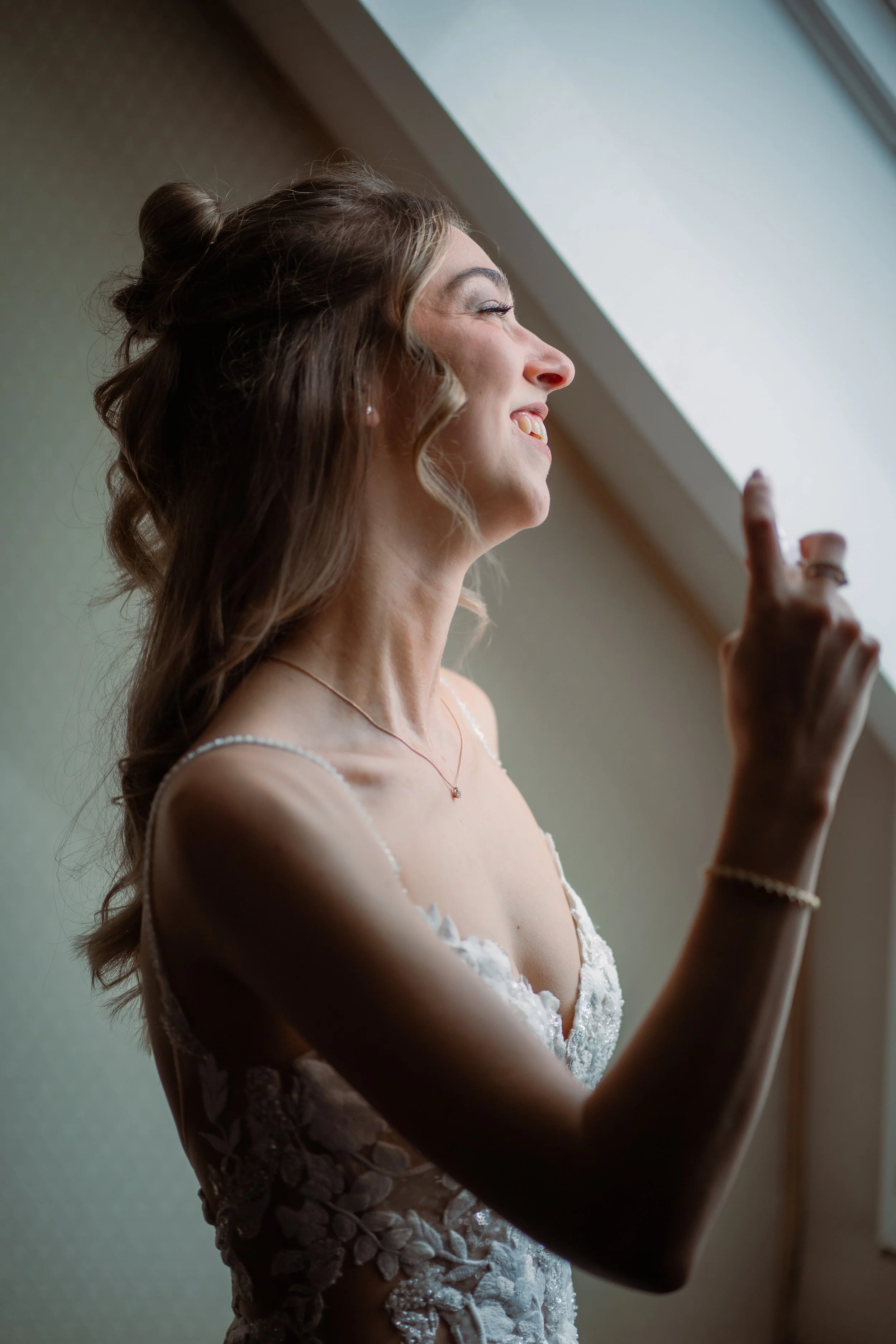 Braut in weißem Hochzeitskleid, lächelnd und in der Nähe eines Fensters, während sie nach draußen schaut.