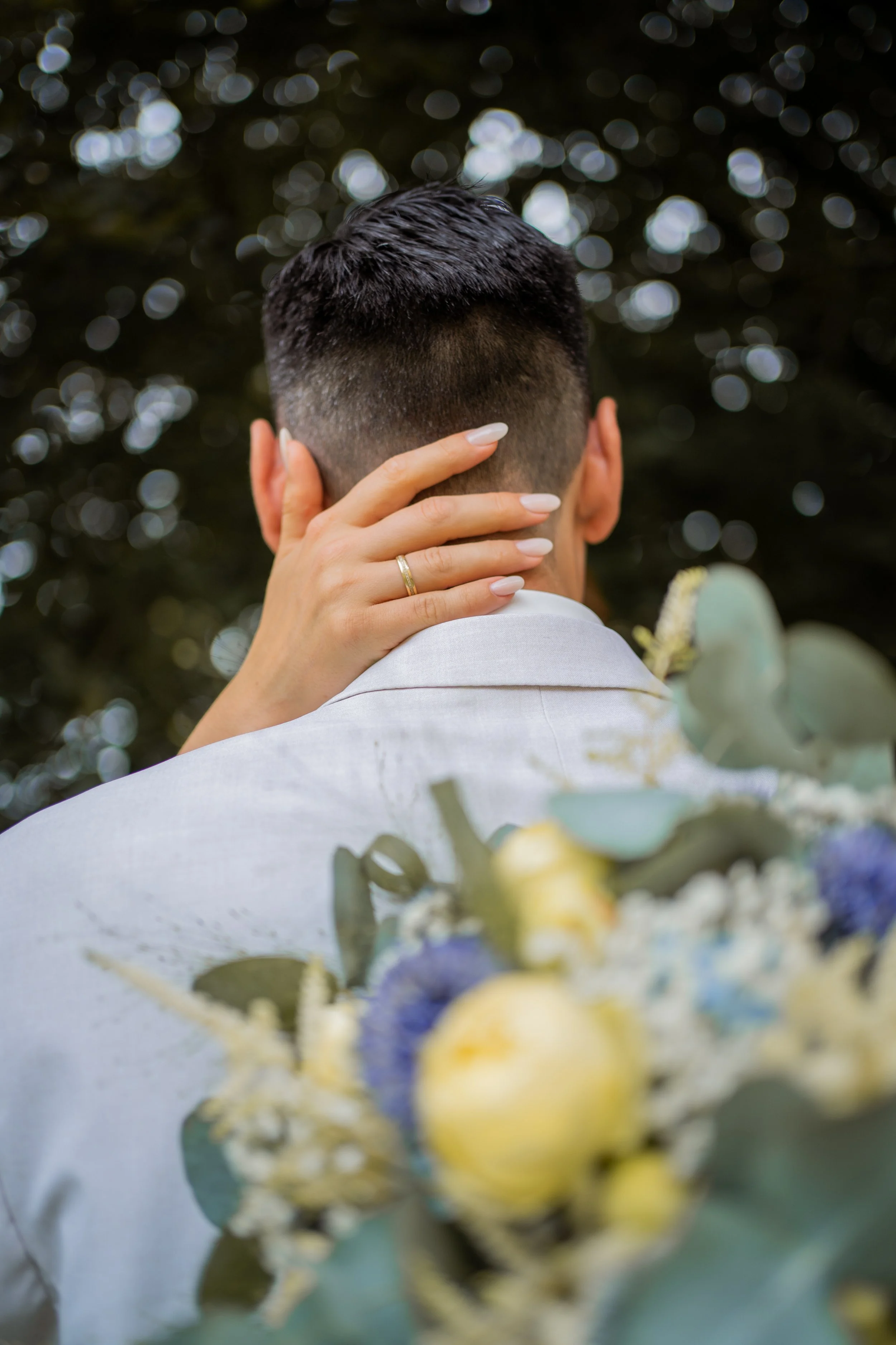 Nahaufnahme eines Mannes, der von hinten zu sehen ist, mit kurzer dunkler Frisur und Hand im Nacken, im Hintergrund unscharfe Lichter. Im Vordergrund unscharfer Blumenstrauß, hauptsächlich in Blau, Gelb und Grün.