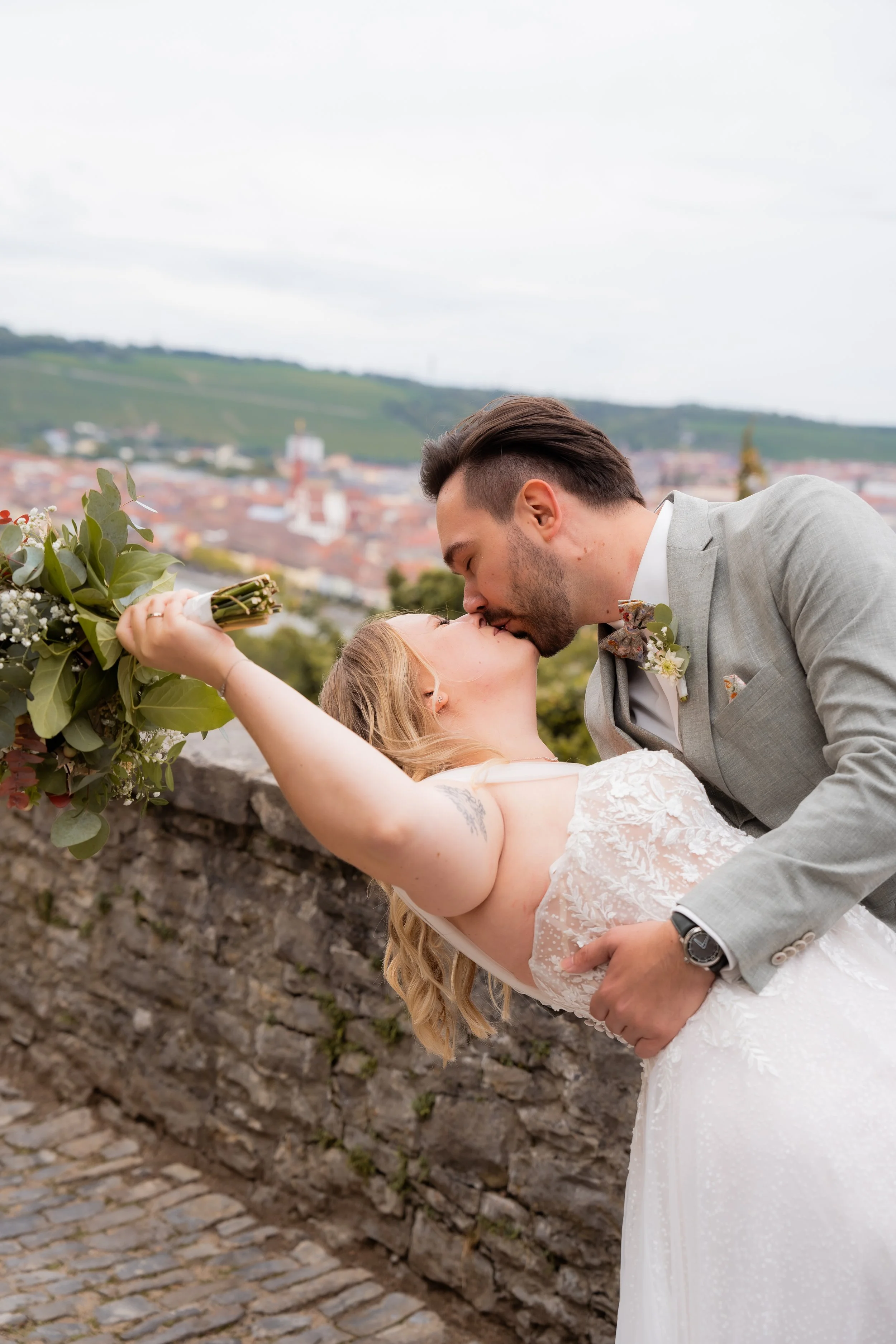 Ein frisch vermähltes Paar teilt einen Kuss in einer romantischen Kulisse mit einem alten Steinmauer und einer Stadt im Hintergrund, die auf einer sanften Hügellandschaft liegt.