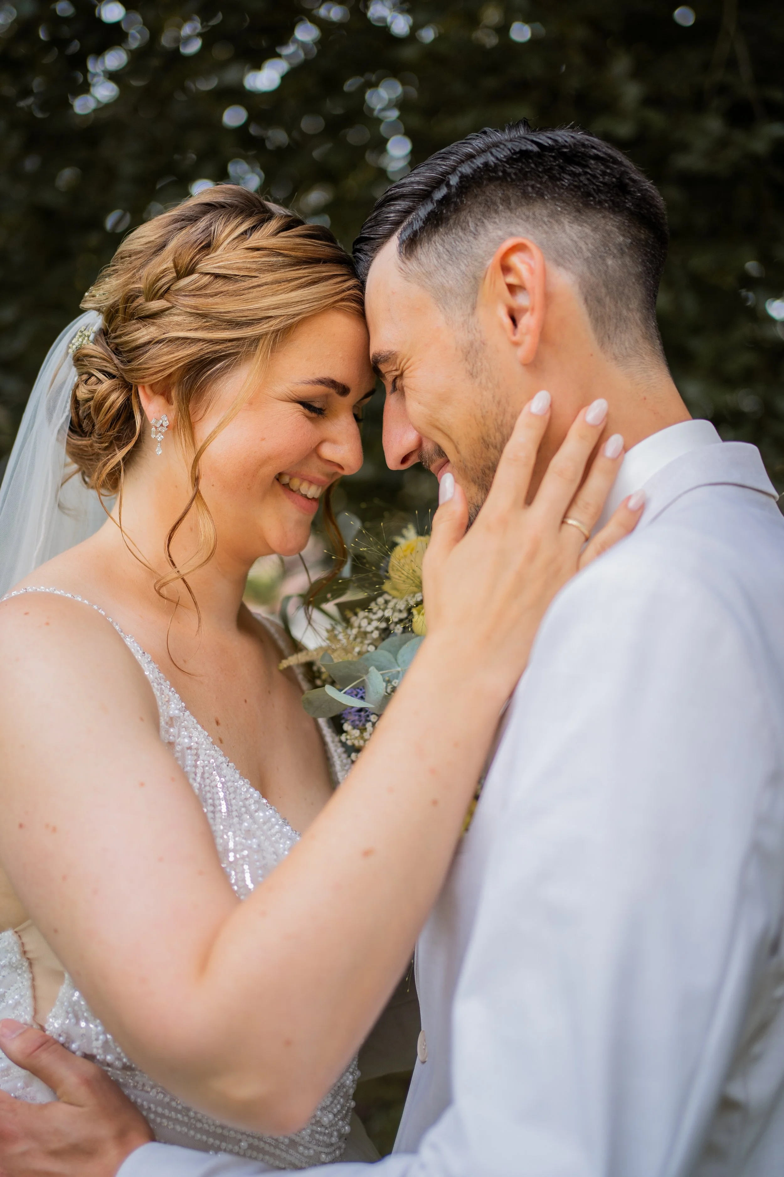 Ein Brautpaar berührt sich mit ihren Stirnen und lächelt. Die Braut trägt ein weißes Hochzeitskleid mit Perlenbesatz und Ohrringe, der Bräutigam trägt einen weißen Anzug.