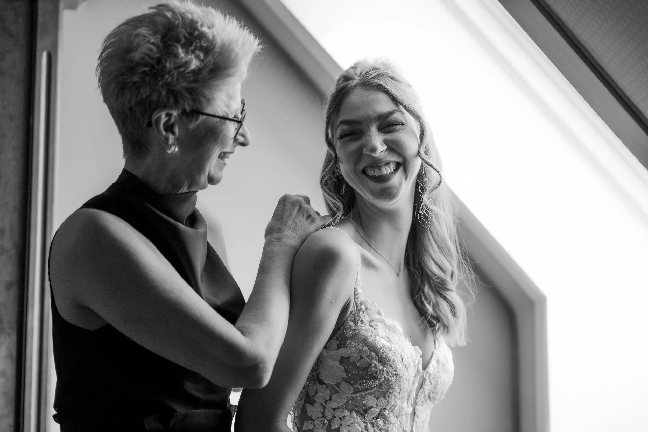 Zwei Frauen lachen und schauen sich an, eine trägt ein Hochzeitskleid, die andere trägt eine Jacke und eine Brille, im Hintergrund eine helle Lichtquelle.
