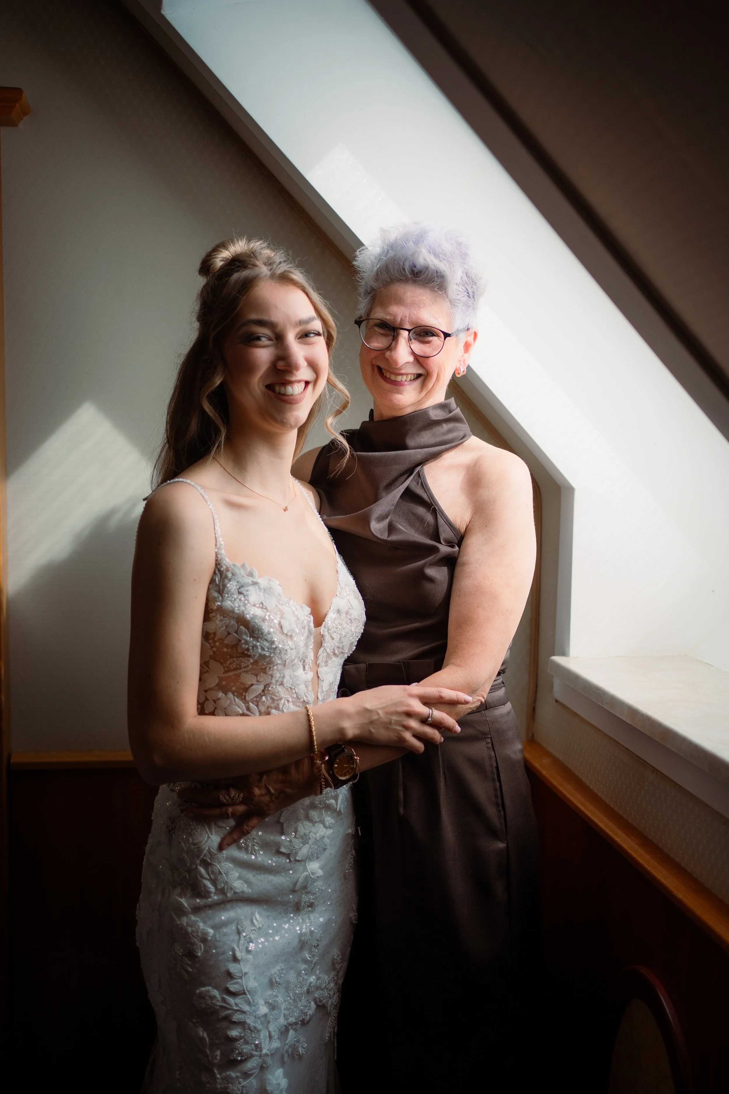 Zwei Frauen, eine jüngere in Brautkleid und eine ältere in eleganter Kleidung, stehen nebeneinander im Raum, lachen und posieren für ein Foto, mit einem Dachfenster im Hintergrund.