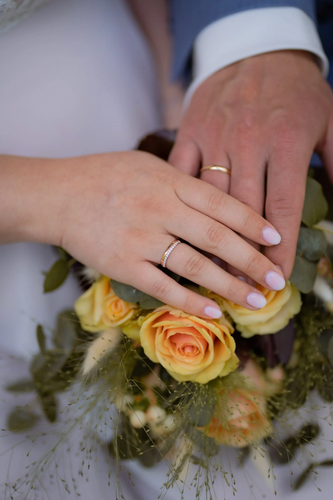 Hände zweier Menschen mit Hochzeitsringen, gehalten über einem Brautstrauß aus gelben und orangen Rosen, umgeben von grünen Blättern und Blumen