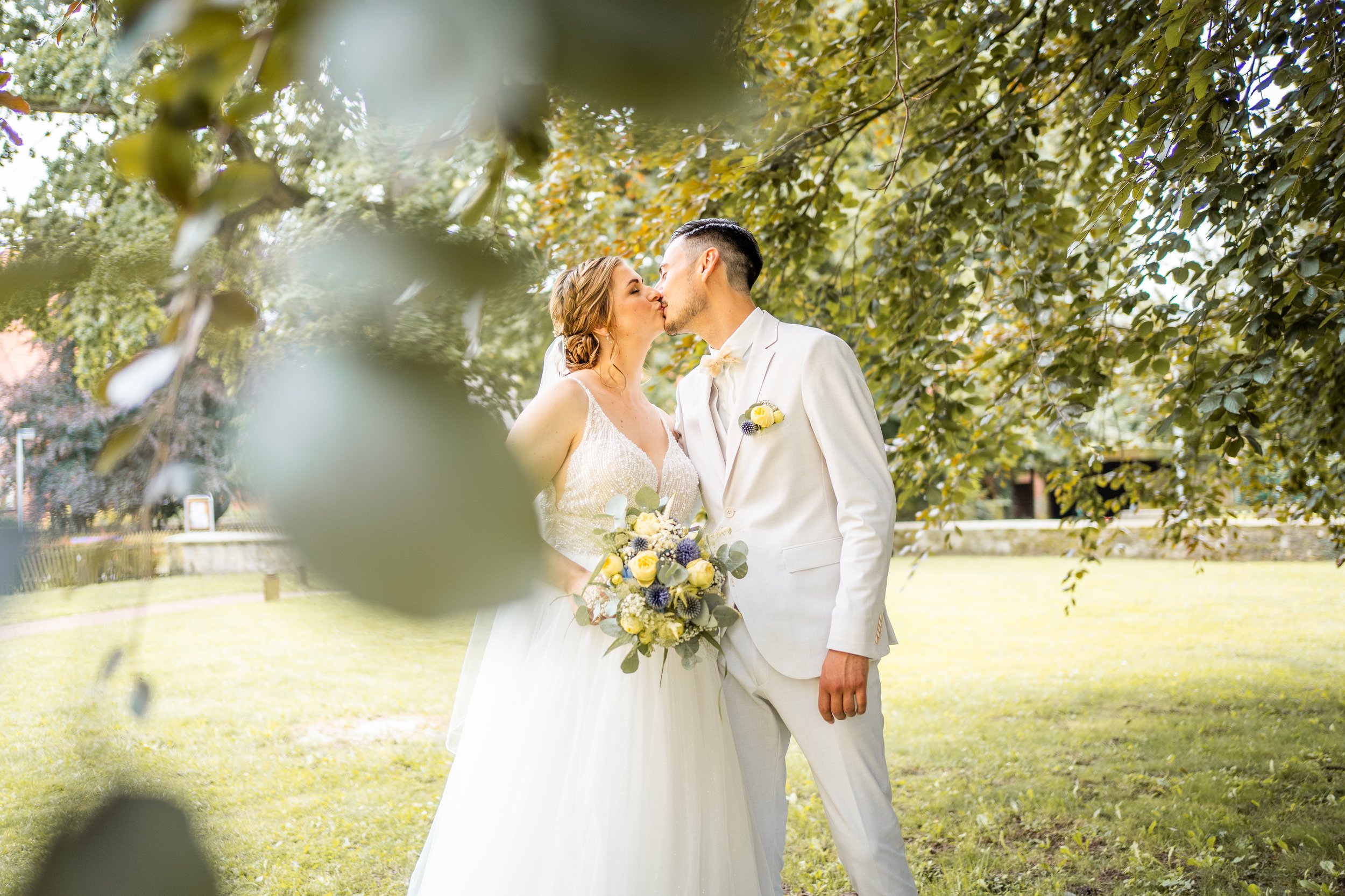 Ein frisch vermähltes Paar beim Kuss im Freien, umgeben von grünen Blättern und Bäumen bei sonnigem Wetter.