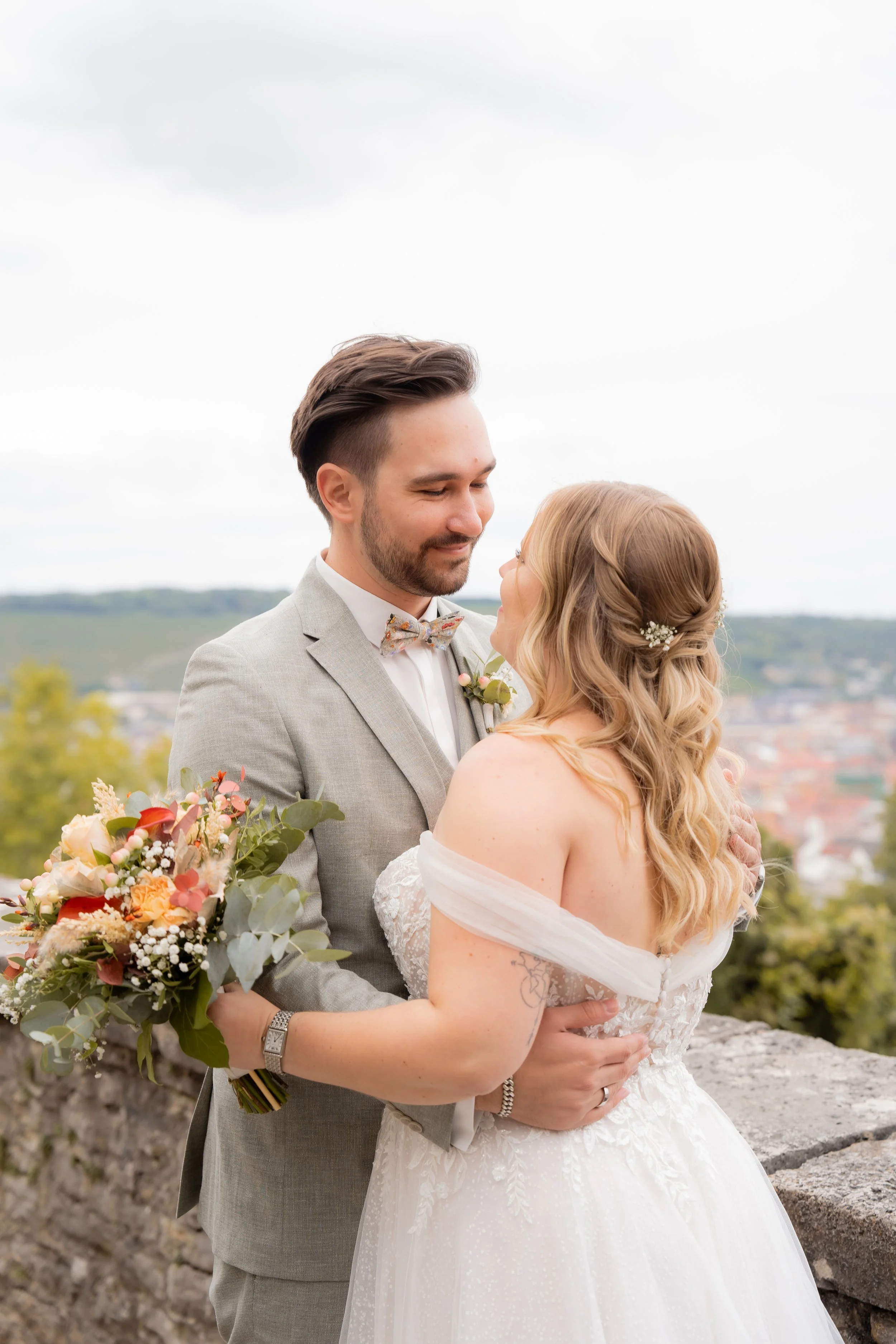 Ein Brautpaar umarmt sich bei ihrer Hochzeit im Freien, mit Blick auf eine Stadt im Hintergrund, die Braut trägt ein weißes Kleid mit Spitze und das Brautkleid ist an den Schultern offen, der Bräutigam trägt einen hellgrauen Anzug mit Fliege und hat 