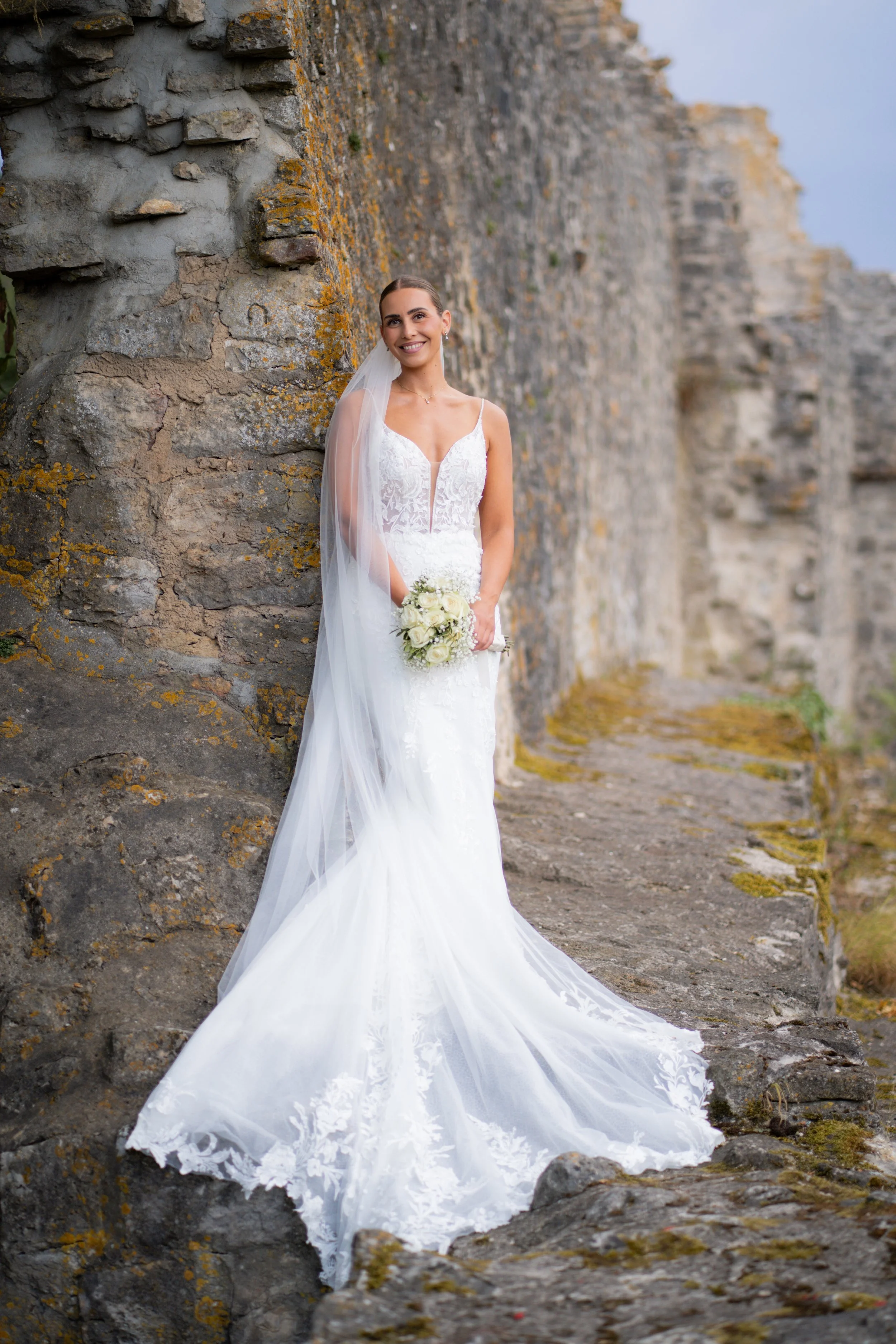 Braut in weißem Brautkleid mit Blumenstrauß vor einer alten Steinmauer