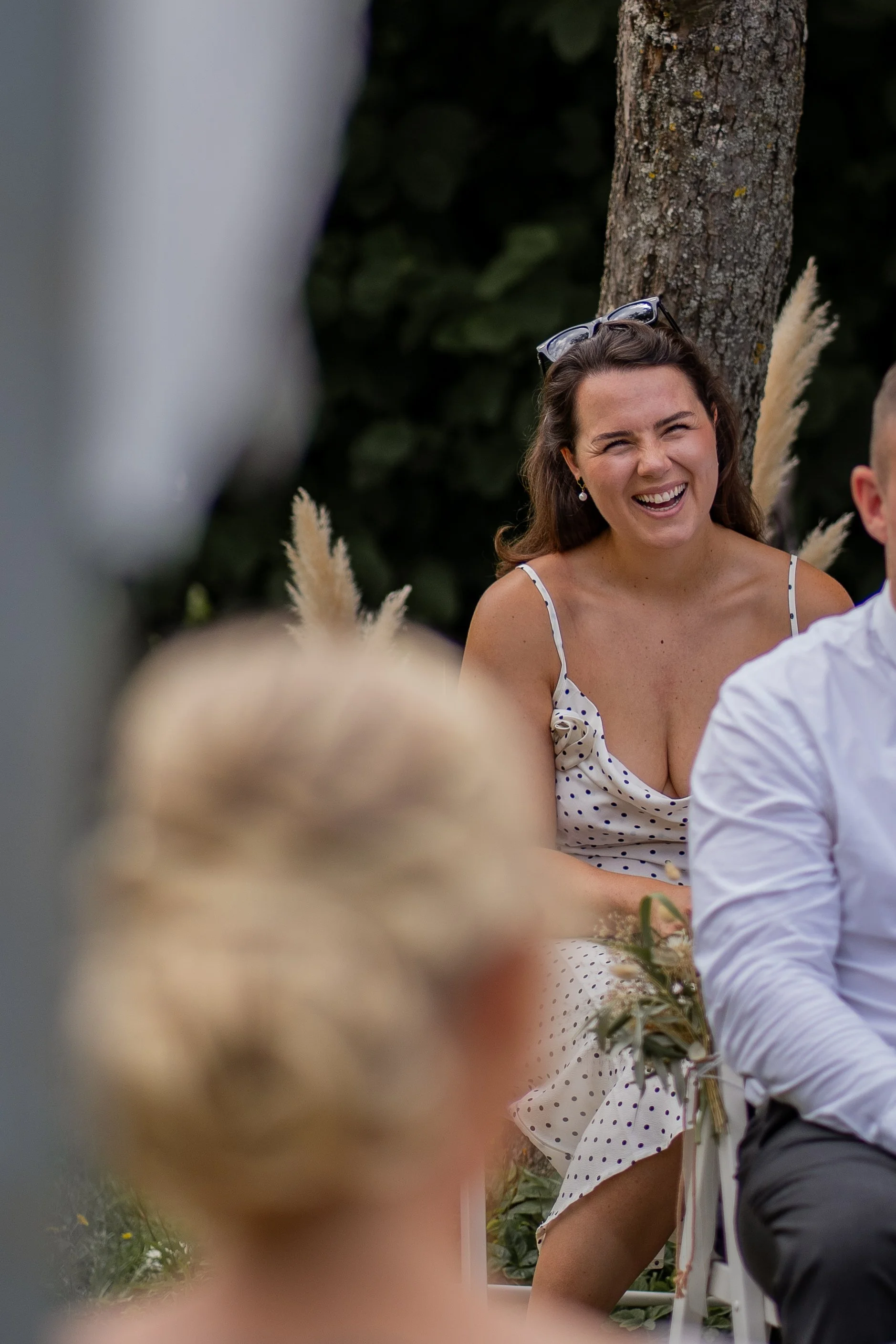 Eine lachende Frau im weißen Kleid mit schwarzen Punkten bei einer Hochzeit im Freien, umgeben von Pflanzen und dekorativen Pampasgräsern.