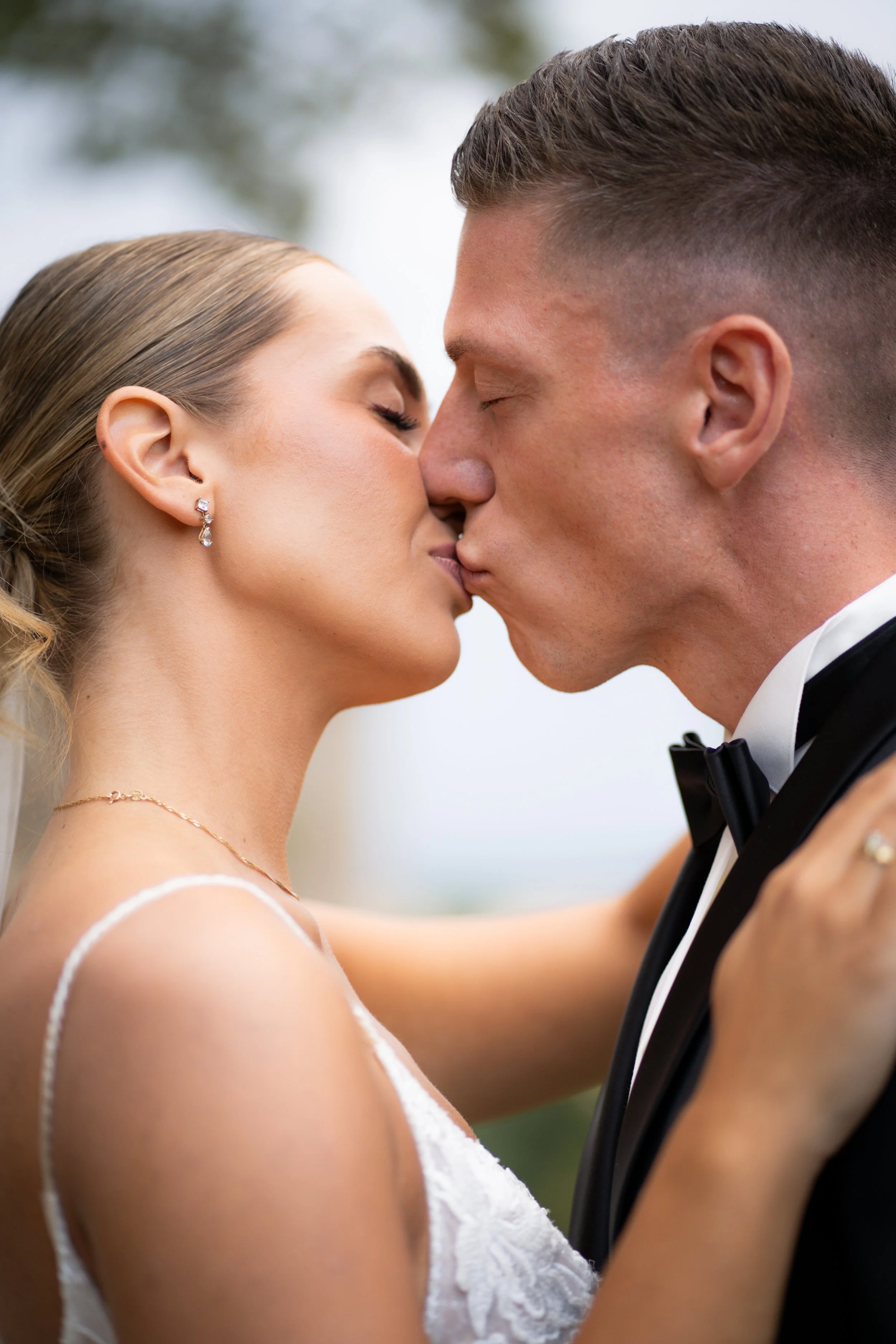 Ein Brautpaar küsst sich bei ihrer Hochzeit, sie trägt ein weißes Kleid und Ohrringe, er trägt einen schwarzen Anzug mit Fliege.