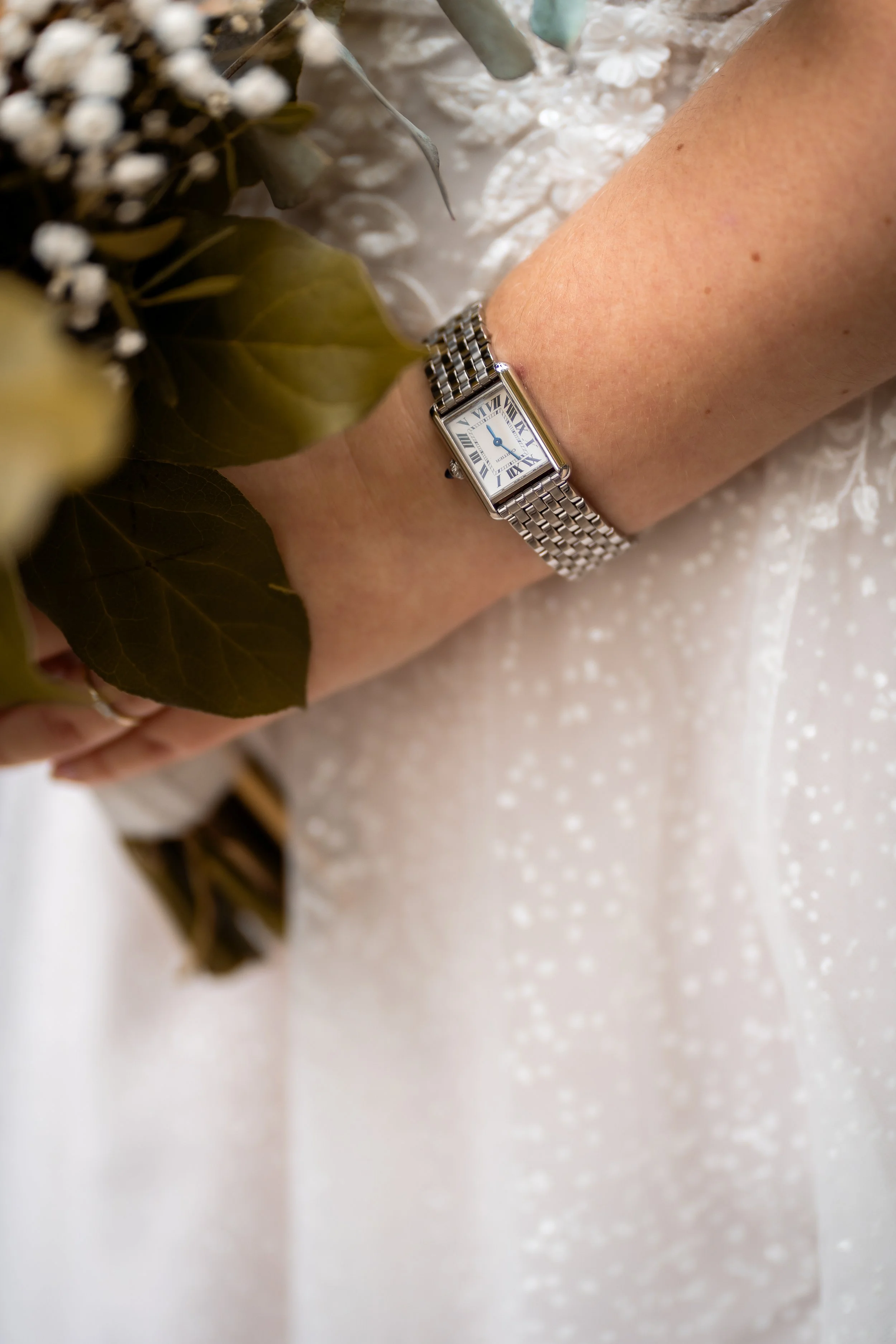 Ein Handgelenk mit einer silbernen Armbanduhr, die römische Ziffern und blaue Zeiger zeigt, liegt auf einem weißen, glitzernden Stoff. Im Hintergrund sind einige grüne Blätter und weiße Blumen.