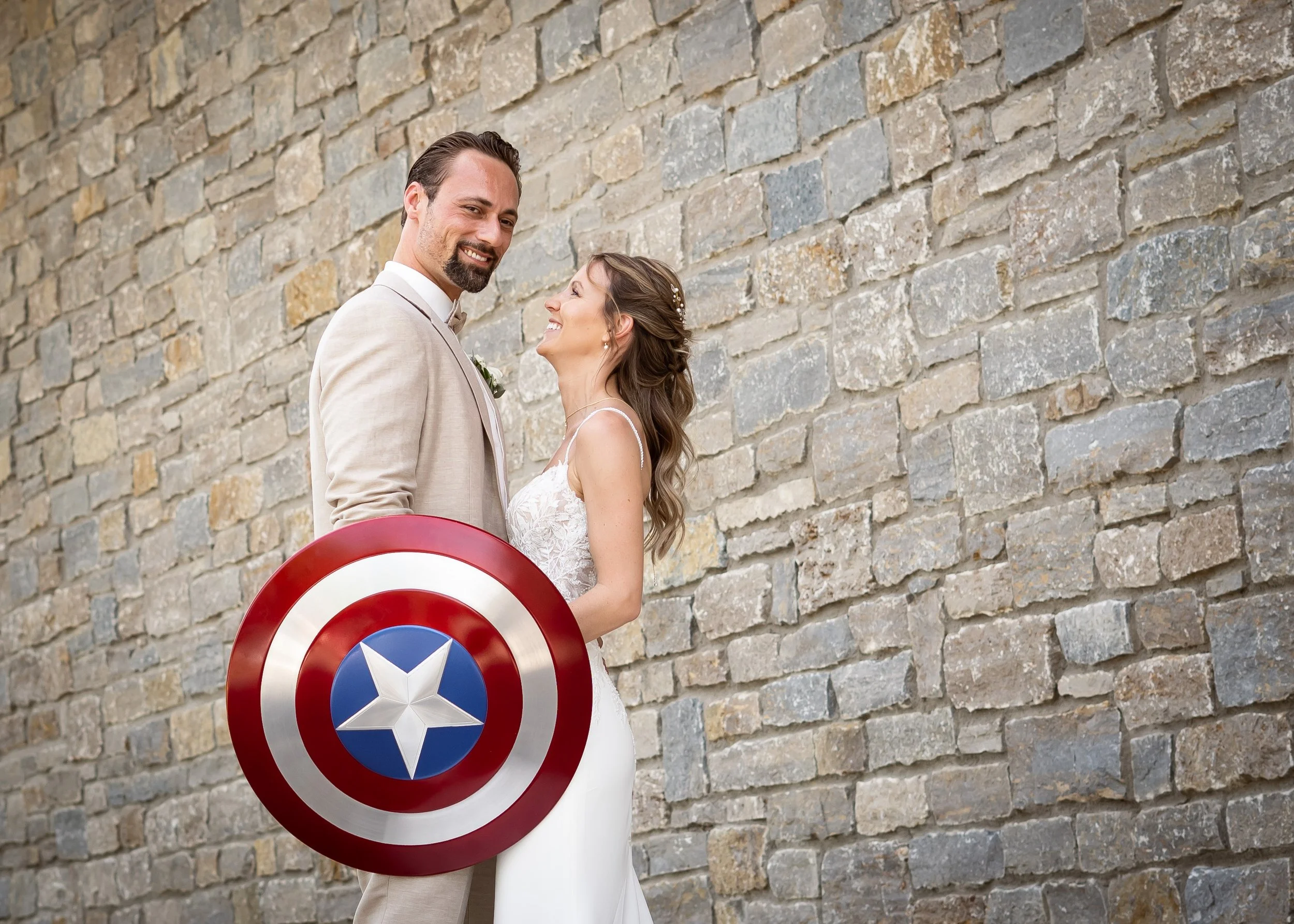 Ein lachendes Brautpaar steht vor einer Steinmauer, die Braut trägt ein weißes Hochzeitskleid, der Bräutigam einen cremefarbenen Anzug. Der Bräutigam hält ein Captain America Schild vor sich, während die Braut ihn lachend ansieht.