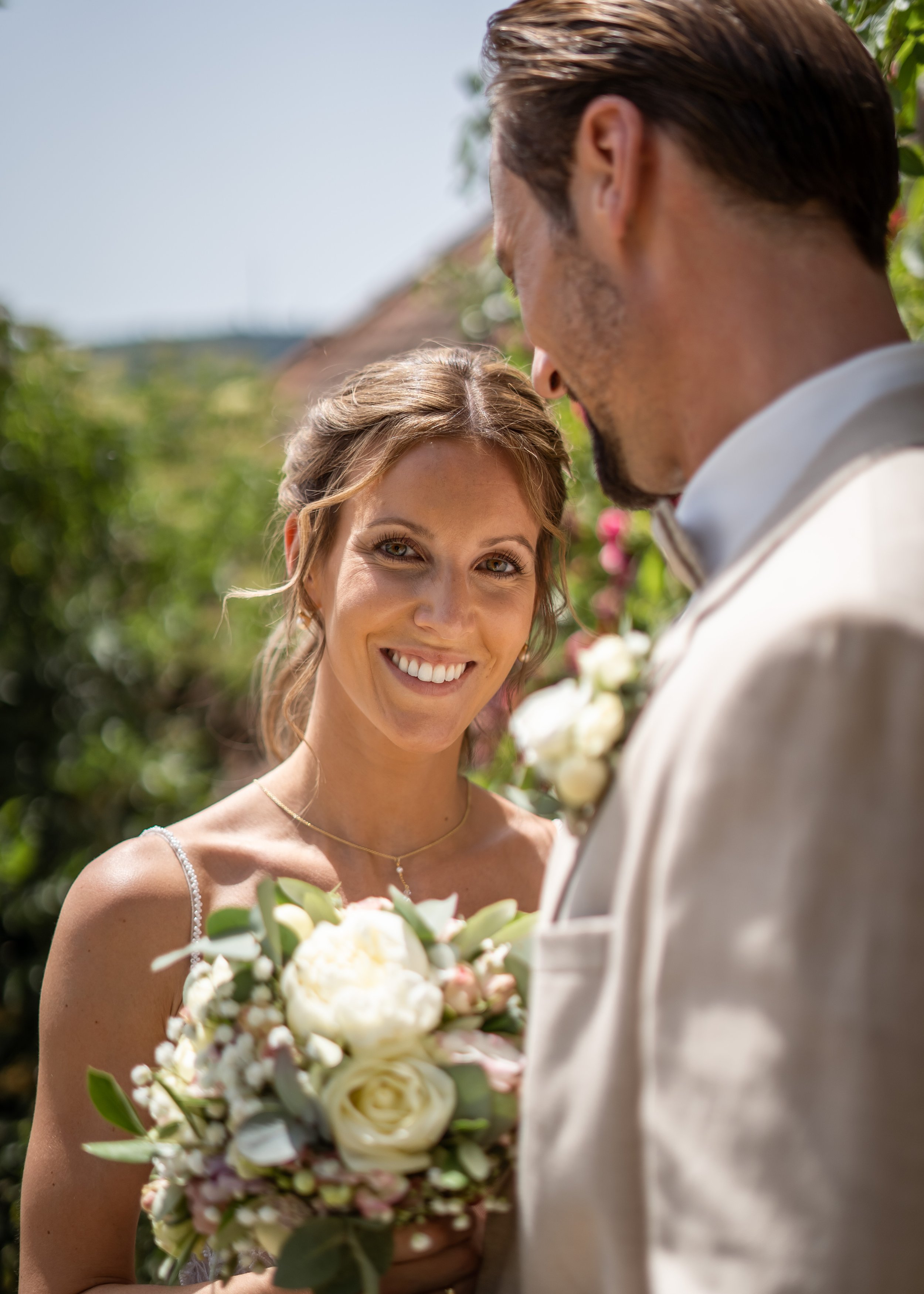 Verliebtes Brautpaar bei einer Hochzeit im Freien, die Frau lächelt freundlich, hält einen Blumenstrauß, während der Mann im Anzug neben ihr steht.