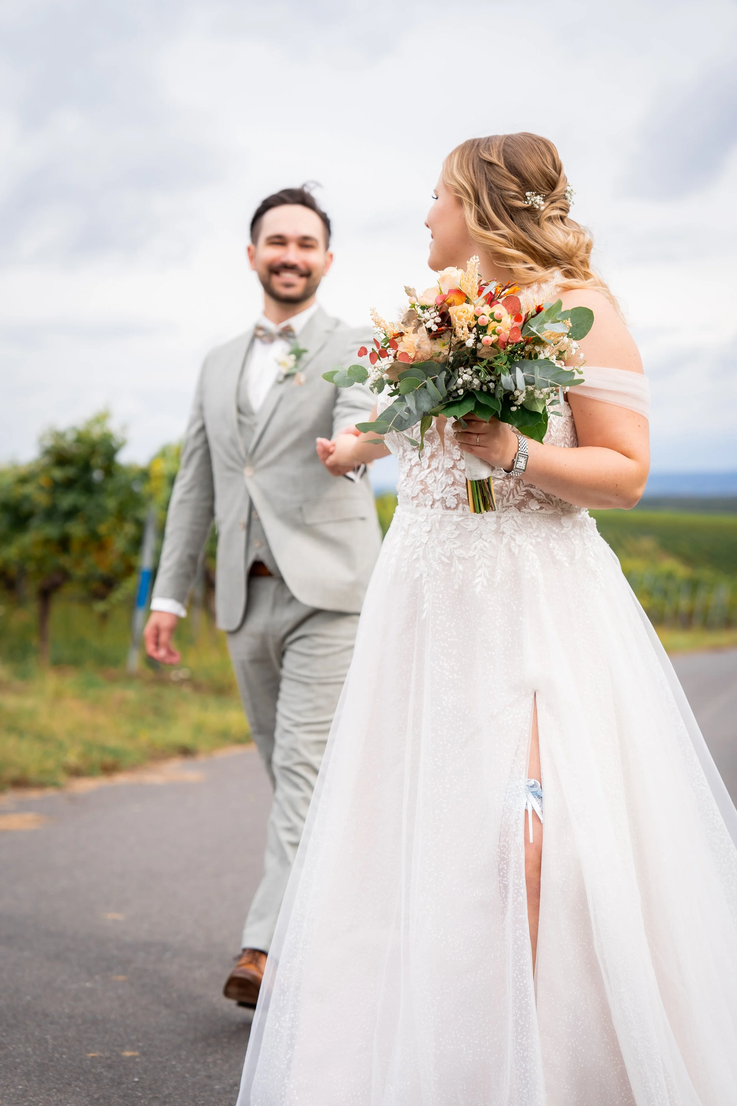 Braut mit Brautstrauß und Brautkleid, Blick zu Bräutigam, im Freien an einem bewölkten Tag.