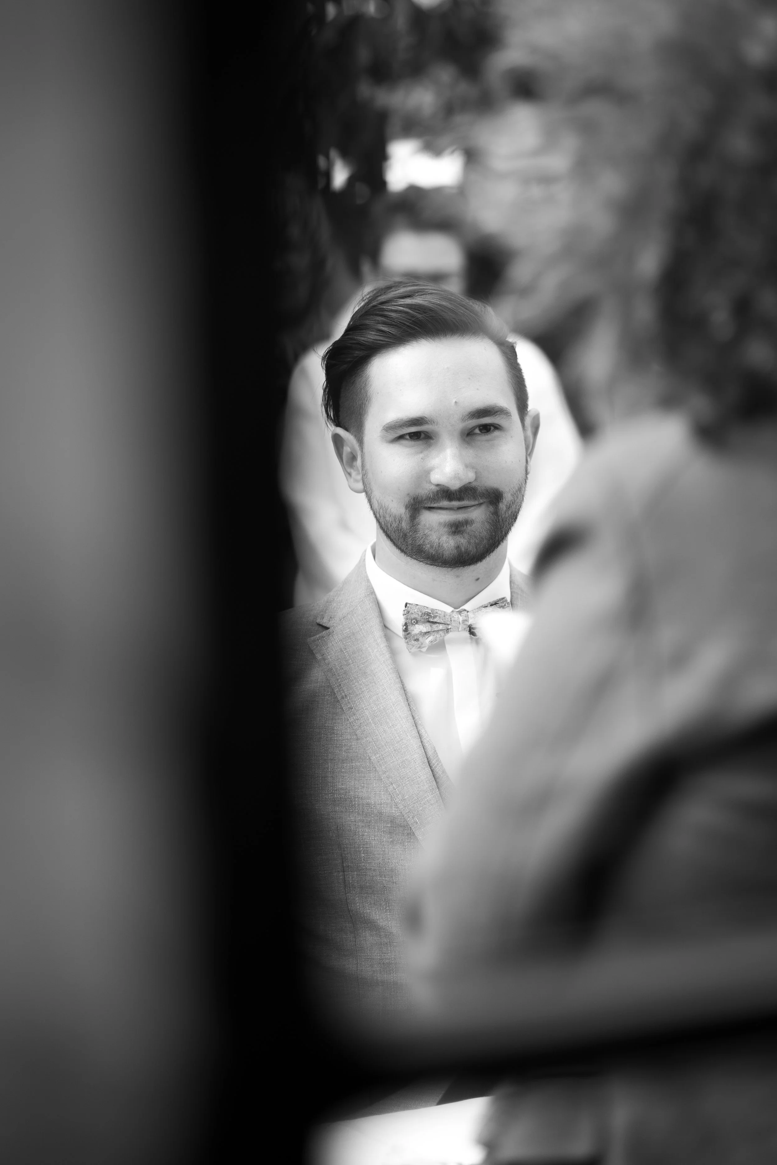 Schwarz-Weiß-Foto eines Mannes mit dunklem, gepflegtem Haar und Bart, der ein formelles Outfit mit Smoking und Fliege trägt. Er blickt leicht lächelnd in die Kamera, im Hintergrund sind unscharfe andere Menschen zu erkennen.