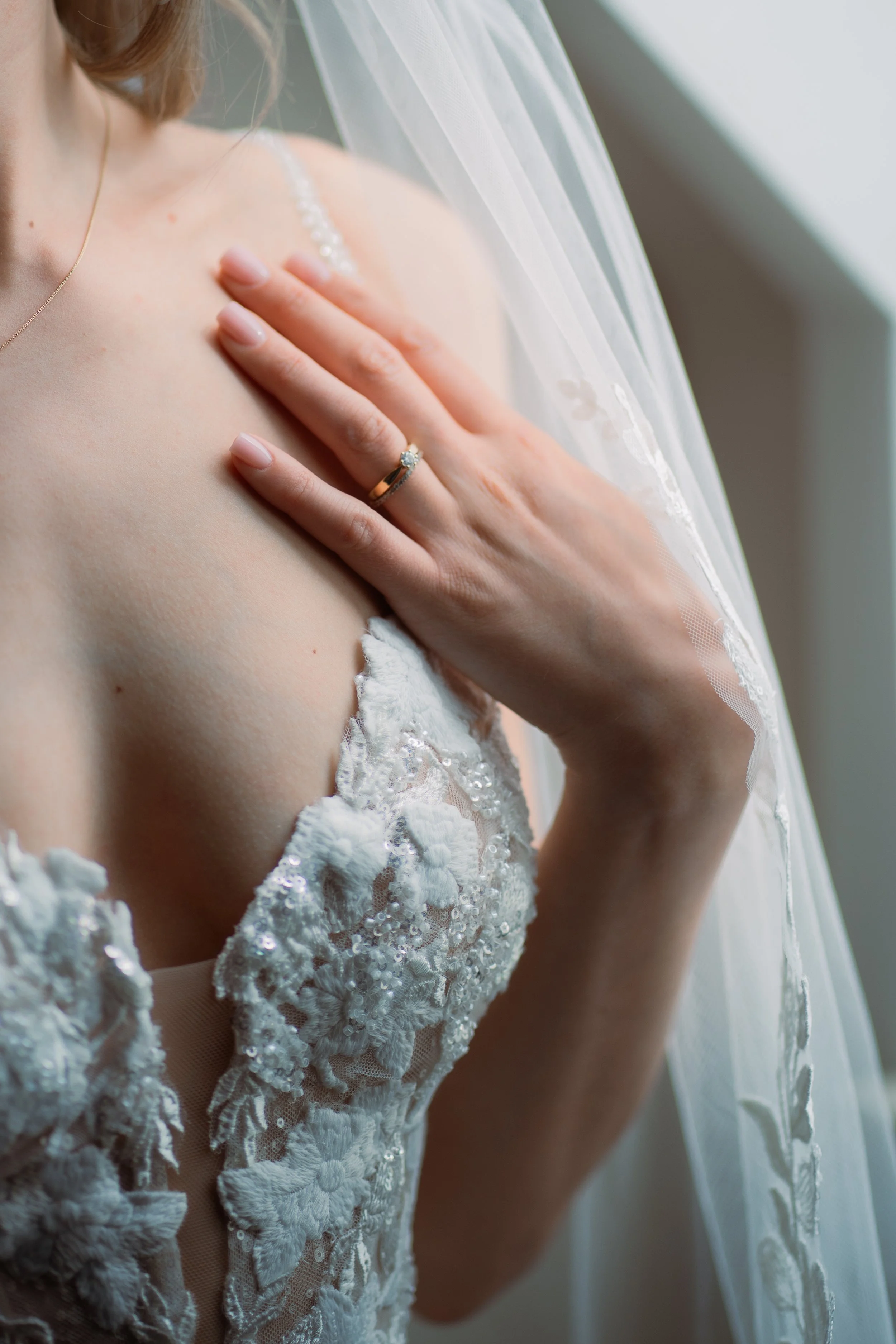 Nahaufnahme der oberen Körperhälfte einer Braut, die ihre Hand auf die Schulter legt, trägt einen Ehering und eine Perlenkette, mit einem weißen Brautkleid mit floraler Stickerei und transparentem Schleier.