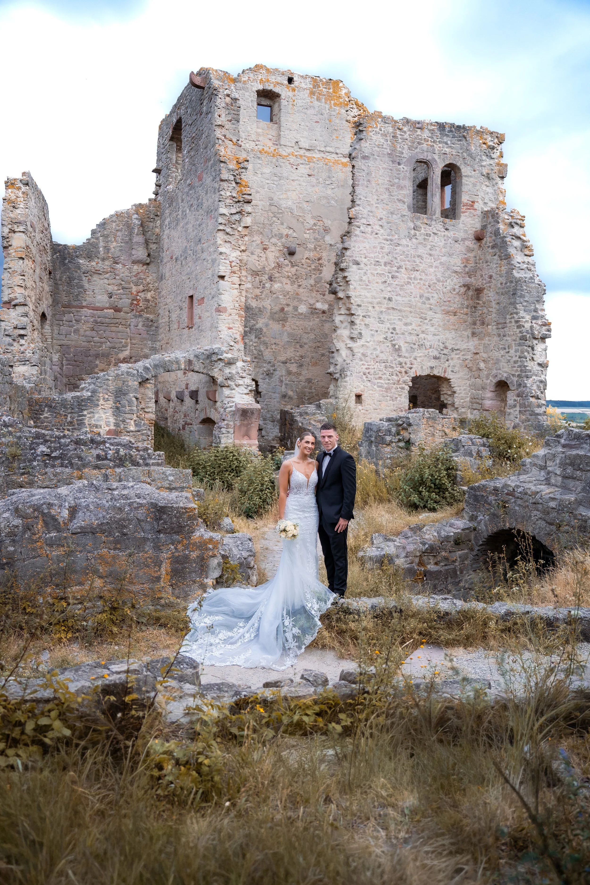 Ein Brautpaar in Hochzeitskleidung vor einer alten, verfallenen Steinburg im Freien.