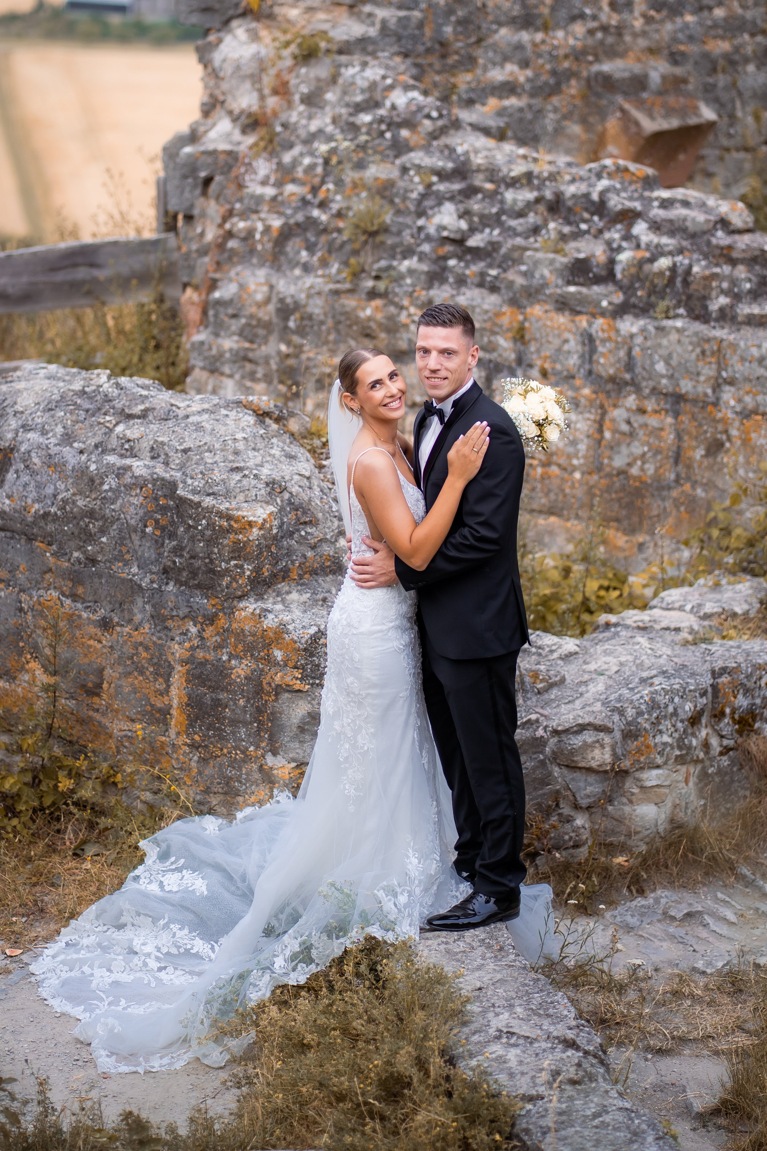 Ein glückliches Brautpaar beim Fotoshooting vor einer alten Steinmauer im Freien, mit der Braut in einem weißen Hochzeitskleid und der Bräutigam in einem schwarzen Anzug, umgeben von Natur und Herbstfarben.