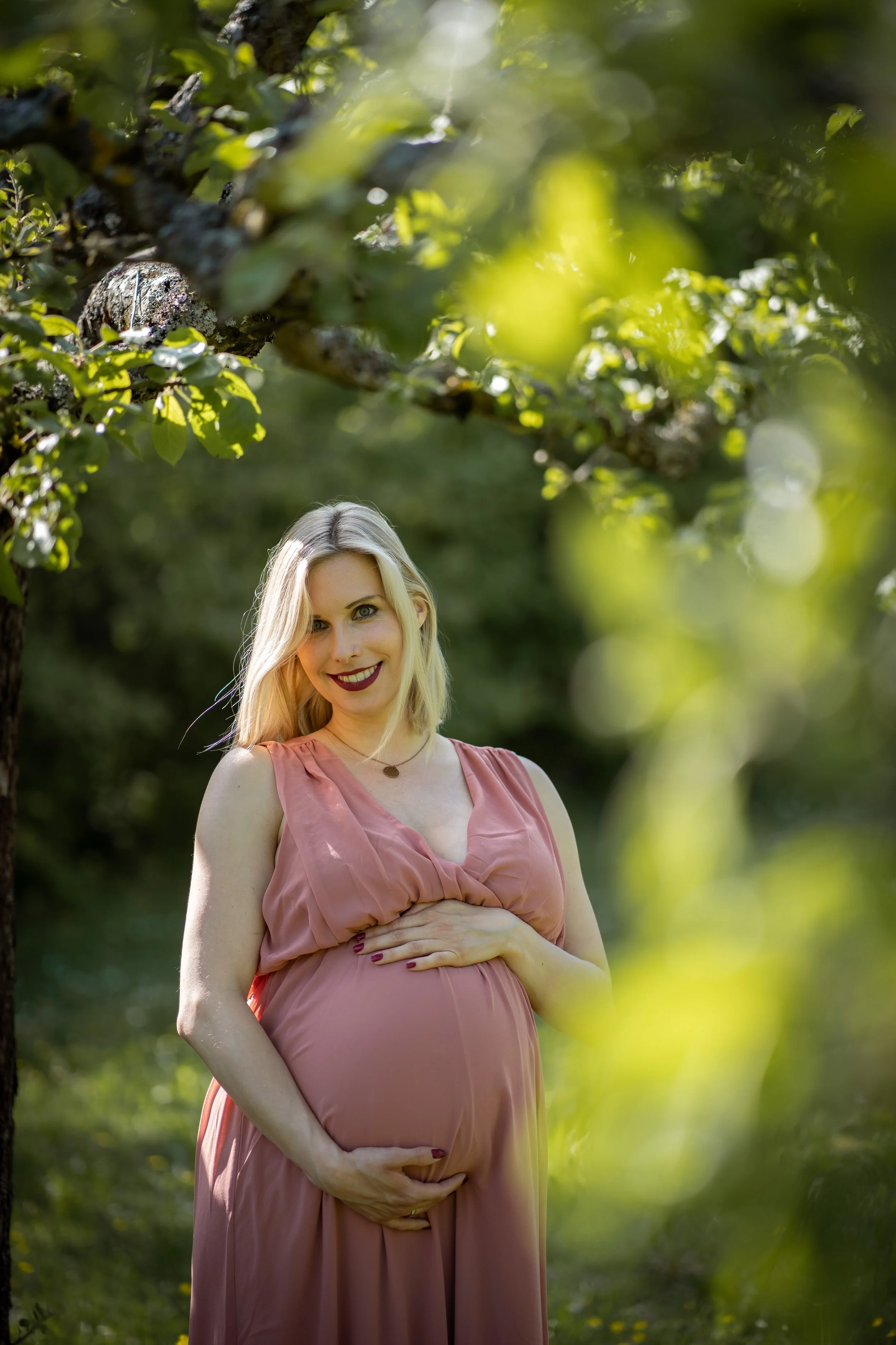 Schwangere Frau posiert auf einer sonnigen Wiese, umgeben von Bäumen und grüner Natur, trägt ein rosa Kleid, lächelt und hält ihre Hand auf den Bauch.