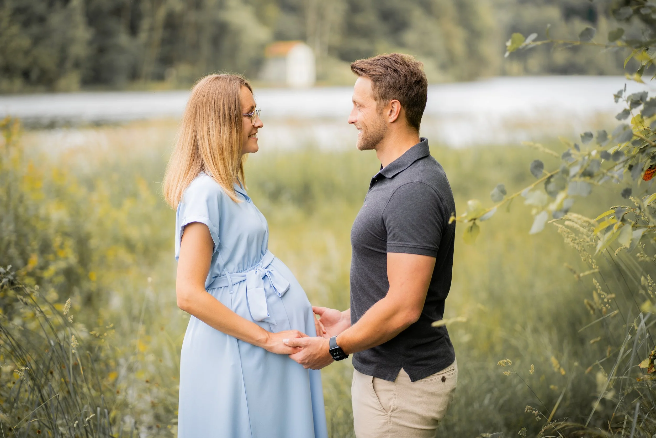 Ein Mann und eine schwangere Frau stehen sich gegenüber, halten Händchen und lächeln sich an, in einer Naturumgebung mit Wasser und Bäumen im Hintergrund.