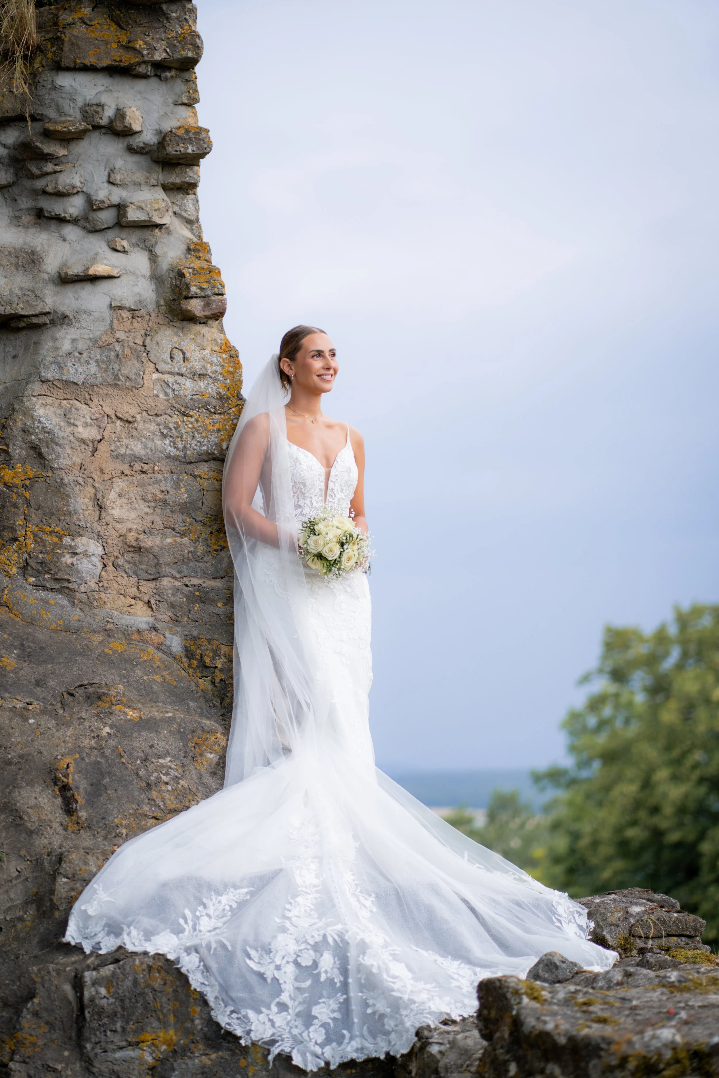 Junge Braut in weißem Hochzeitskleid mit Schleier und Blumenstrauß steht an einer alten Steinmauer mit Blick auf eine grüne Landschaft und bewölkten Himmel.