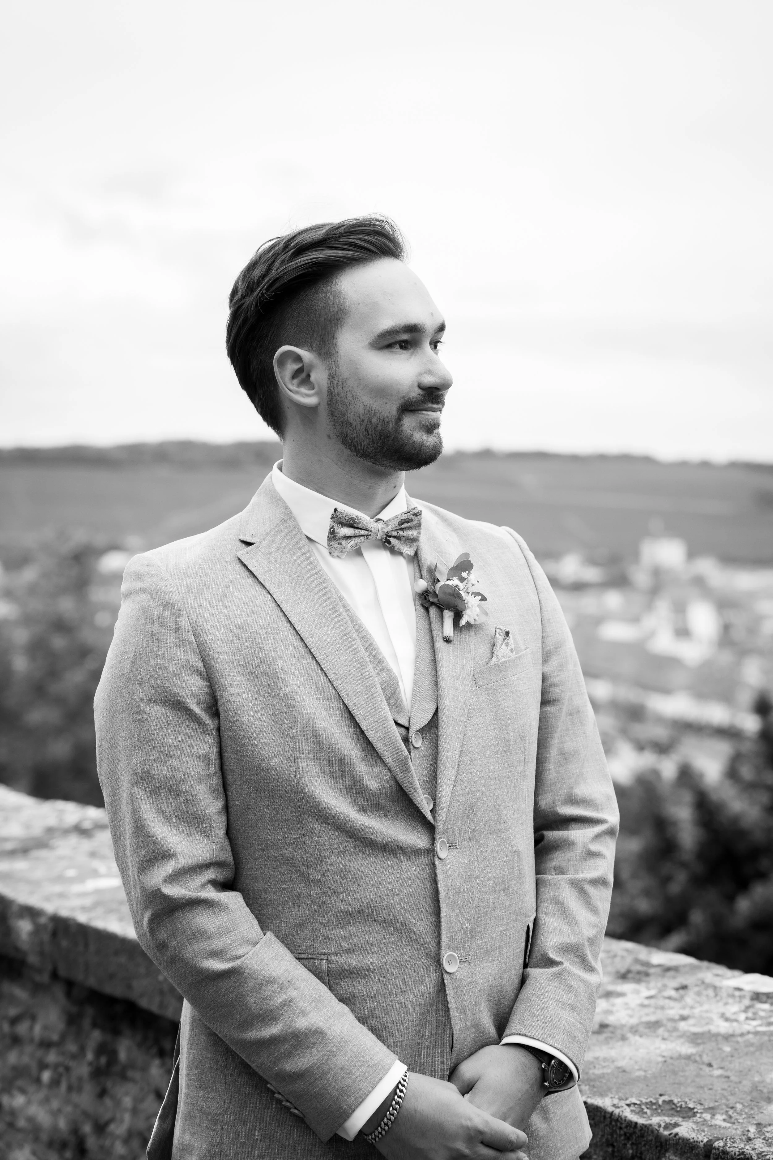 Mann in Anzug mit Blumenboutonnière posiert an einer Mauer im Freien, schwarz-weiß Bild, auf einem Hügel mit Landschaft im Hintergrund.