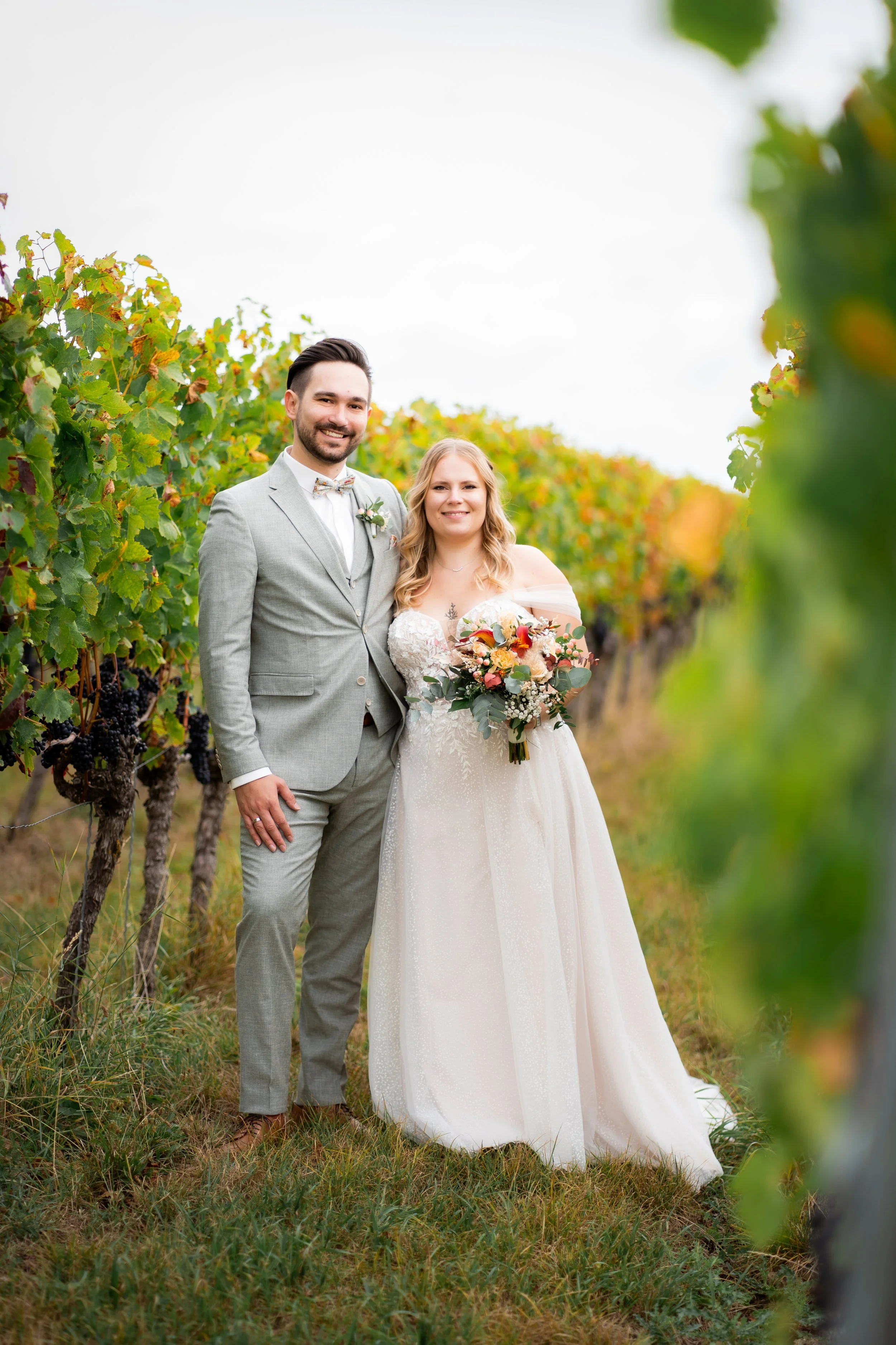 Ein frisch verheiratetes Paar steht im Weinberg, das Brautpaar trägt elegante Kleidung, die Frau hält einen Blumenstrauß, beide lächeln glücklich.