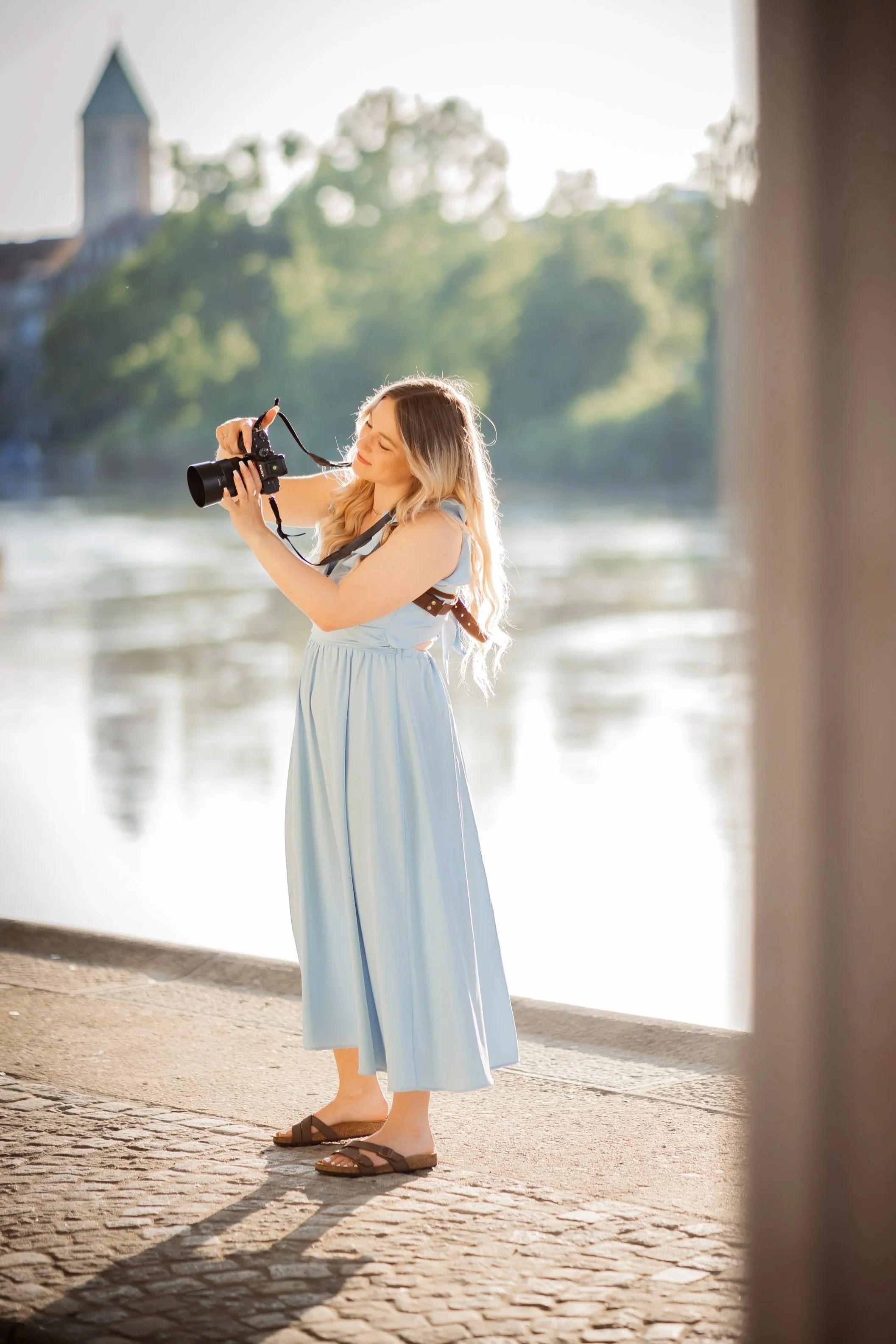 Junge Frau in einem blauen Kleid und Sandalen, die an einem Flussufer steht und eine Kamera hält während sie ein Shooting forografiert.