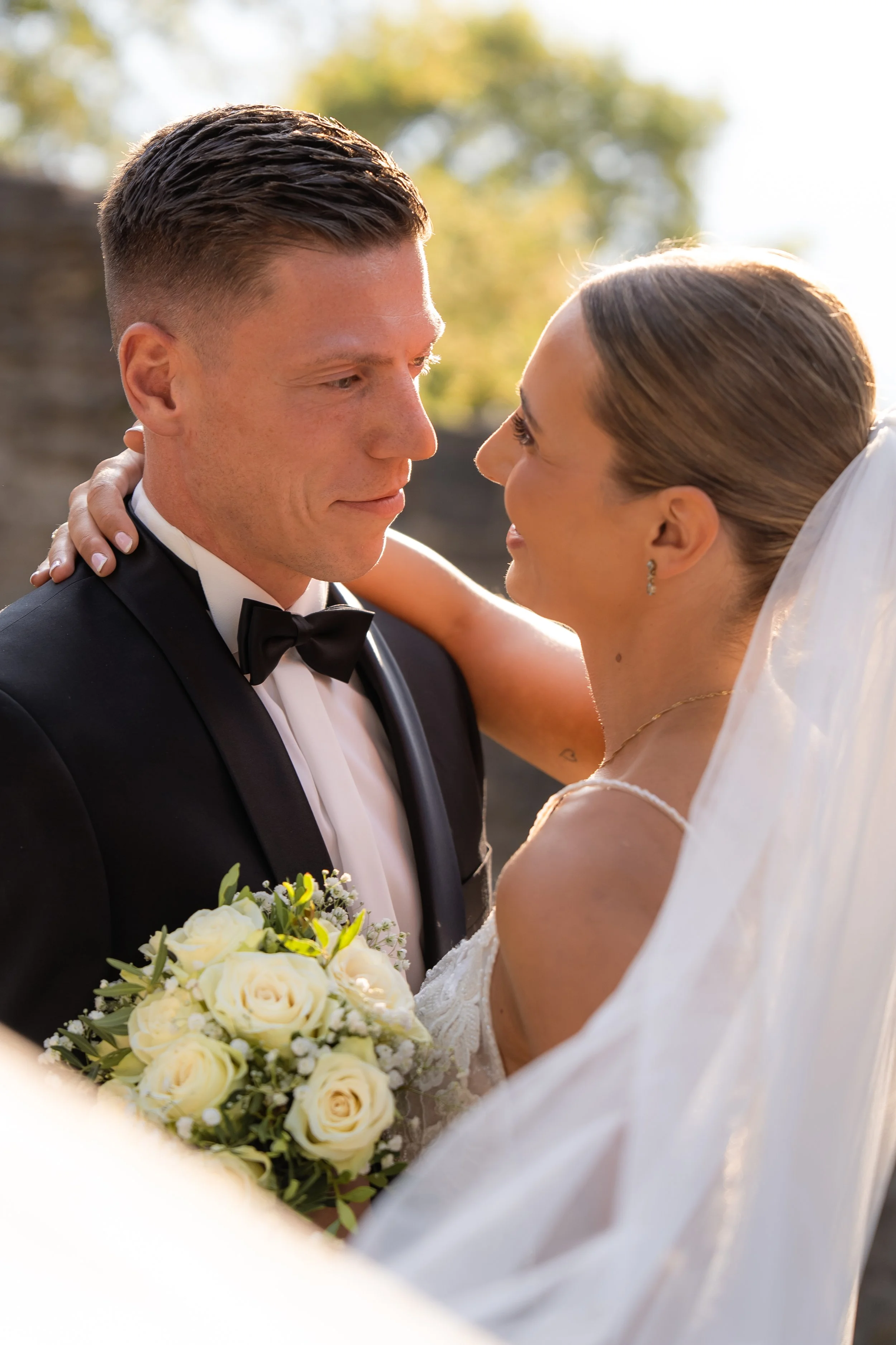 Ein glückliches Brautpaar blickt sich liebevoll an, mit dem Bräutigam in einem schwarzen Anzug und Fliege, und der Braut in einem weißen Kleid mit Schleier, die ein Bouquet aus weißen Rosen hält, bei Sonnenuntergang im Freien.