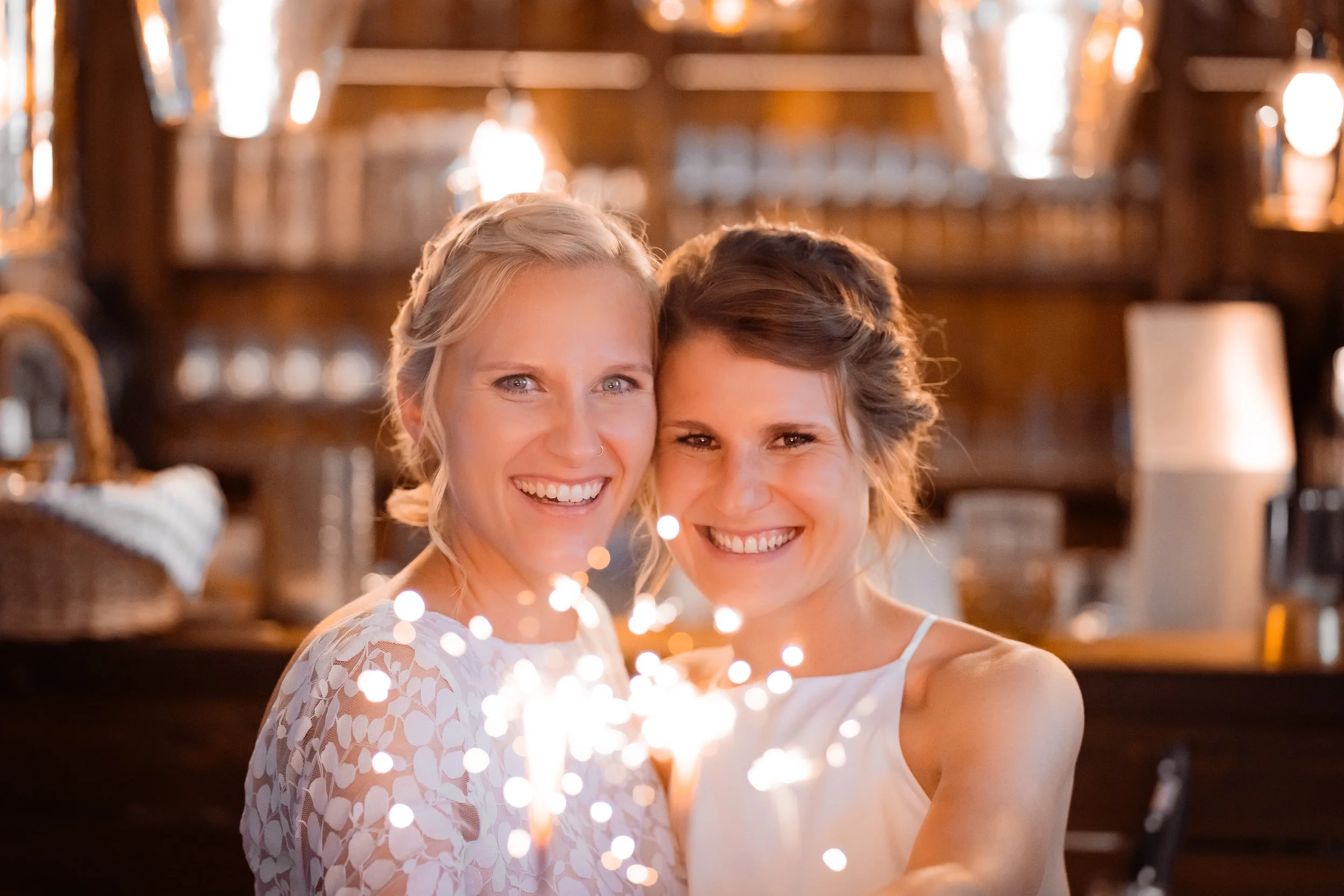 Zwei Frauen lachen und halten leuchtende Wunderkerzen in einer warm beleuchteten, gemütlichen Raum mit Holzmöbeln im Hintergrund.