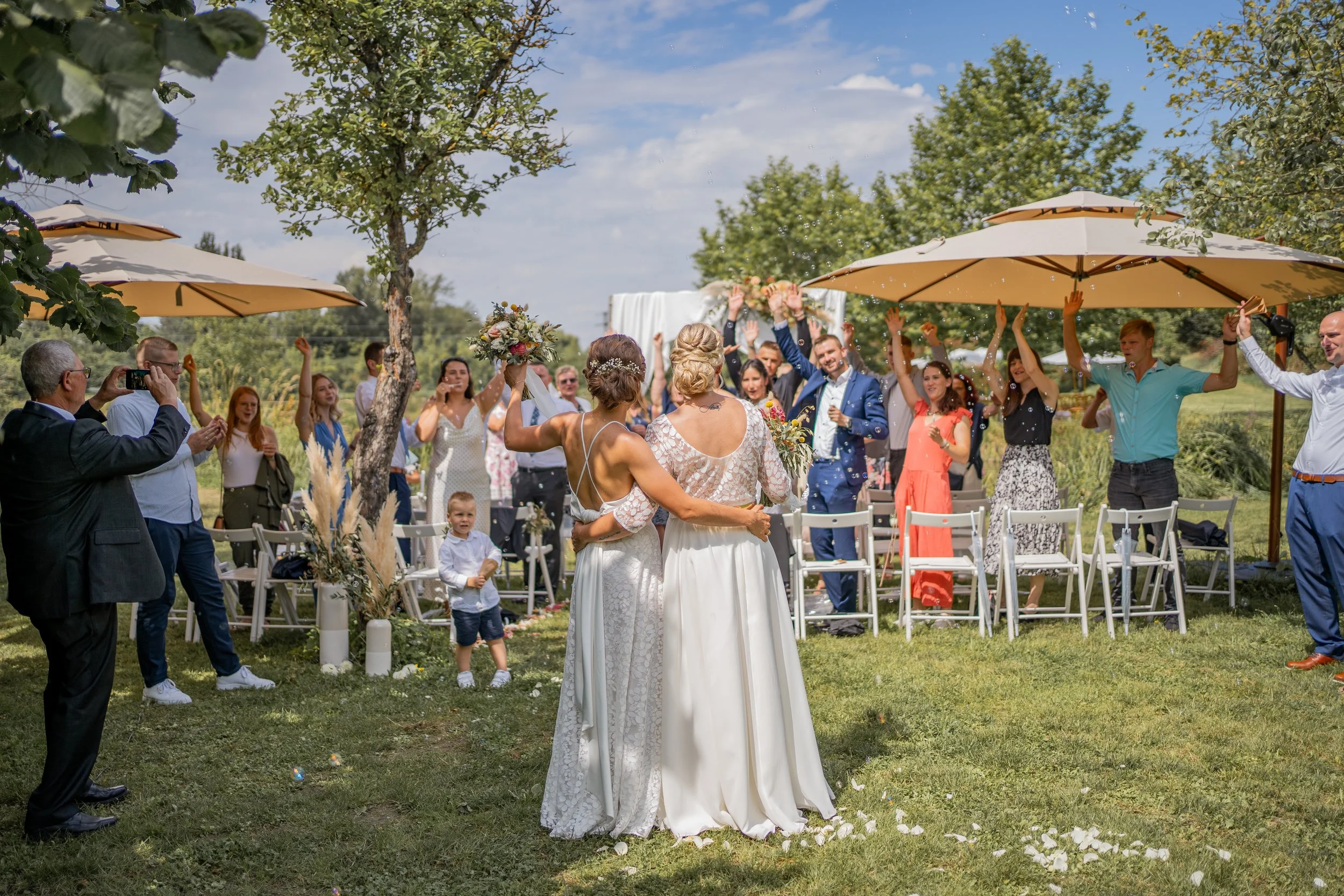 Eine Gruppe von Menschen bei einer Hochzeitszeremonie im Freien, mit zwei Frauen in weißen Brautkleidern im Mittelpunkt, während Gäste unter Sonnenschirmen feiern und sich freuen.