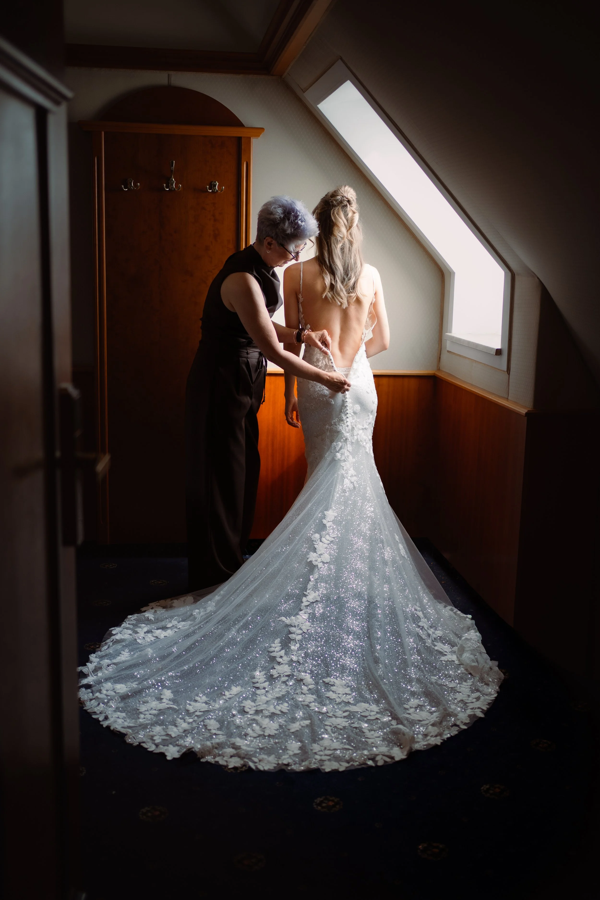 Eine Braut in einem weißen, glitzernden Hochzeitskleid mit Schleppe, die von einer Person, vermutlich einer Schneiderin, im Raum mit Dachfenstern angepasst wird.