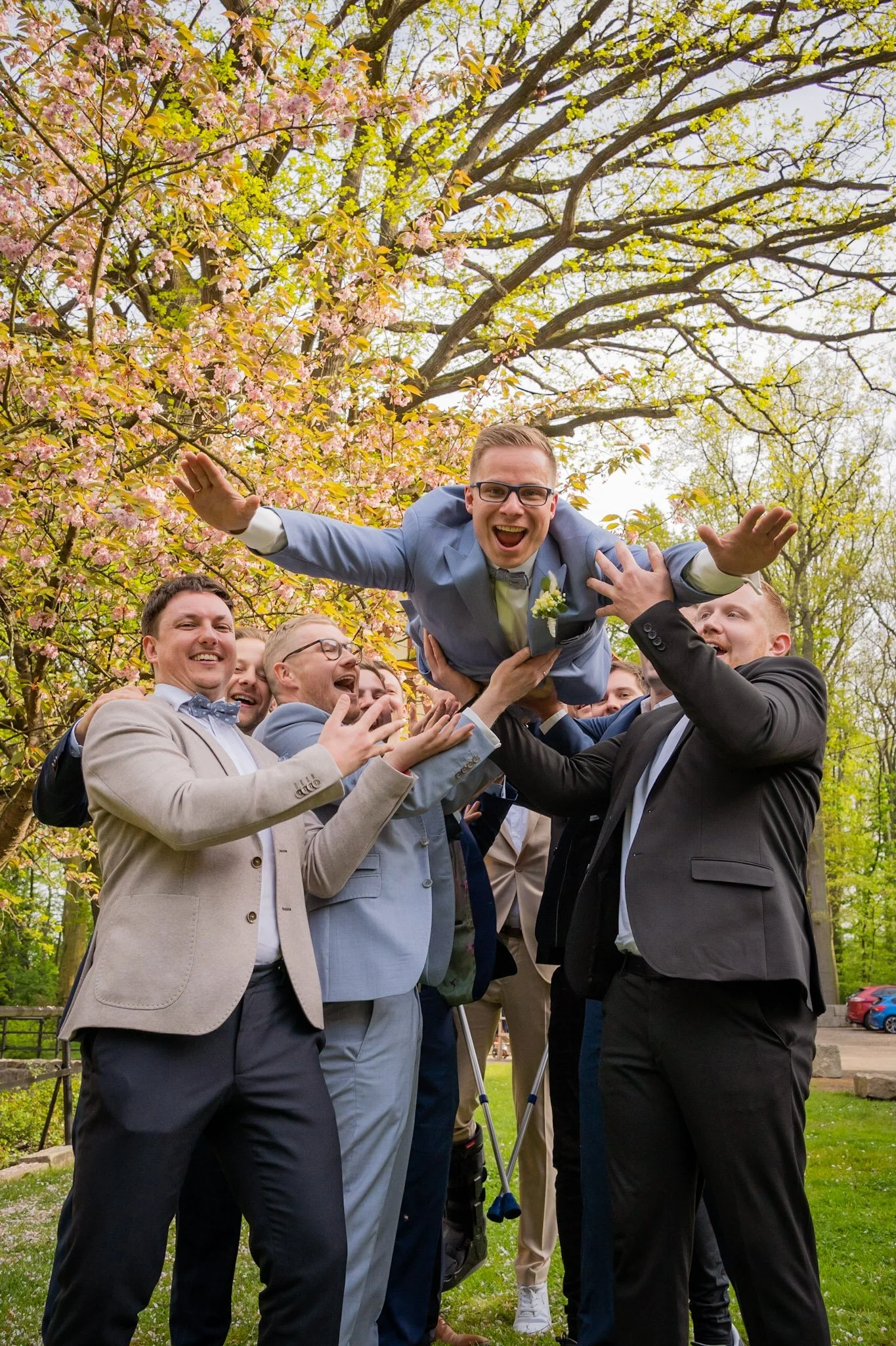 Eine Gruppe von Männern in Anzügen hebt einen jubelnden Mann in die Luft bei einem Outdoor-Event im Park, umgeben von blühenden Bäumen.