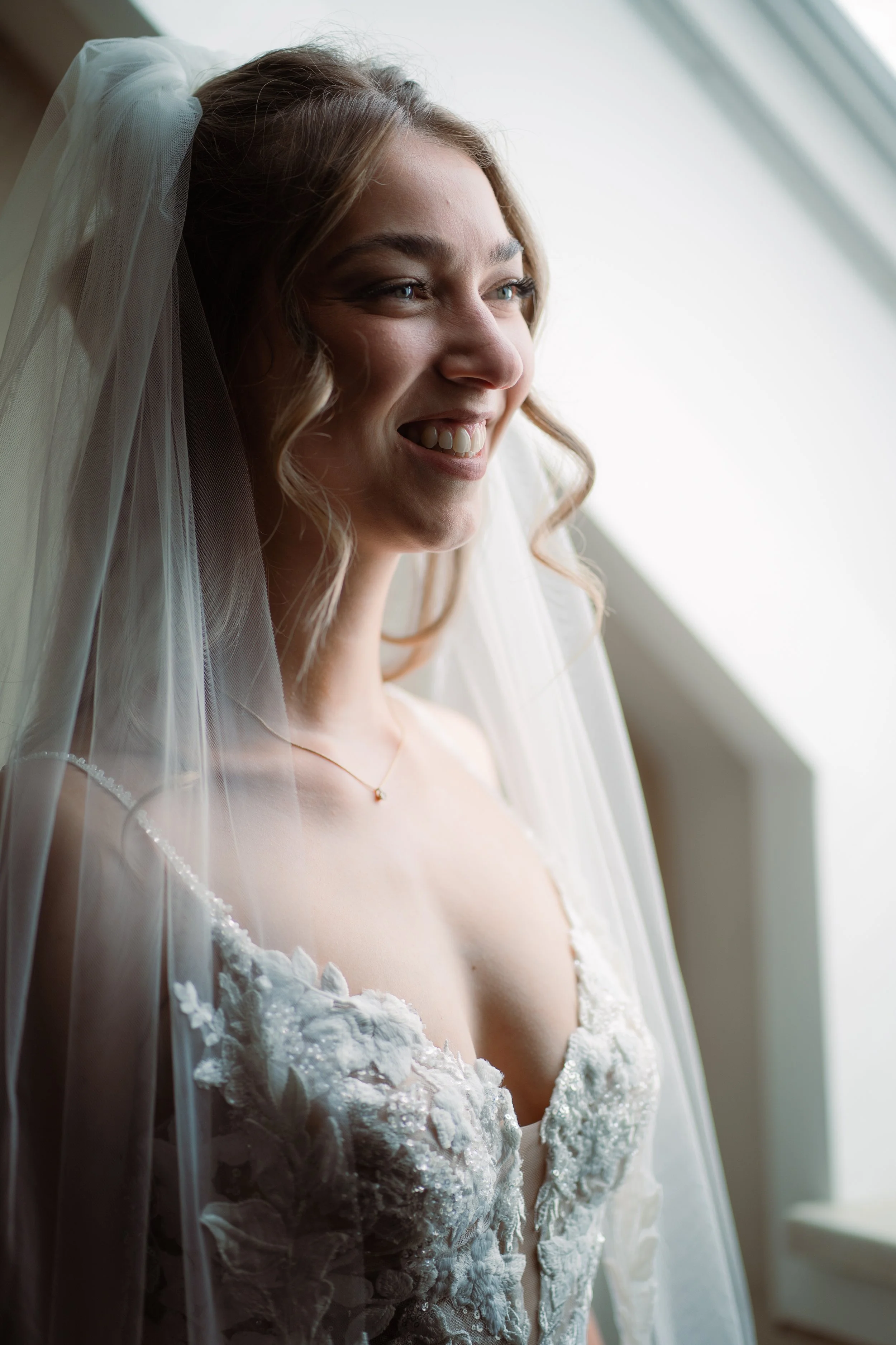 Eine junge Frau, vermutlich eine Braut, trägt ein weißes Brautkleid mit floralen Applikationen und eine Tüll-Voile. Sie hat lange, lockige Haare und ein freundliches Lächeln, während sie nach draußen schaut, bei natürlichem Licht.