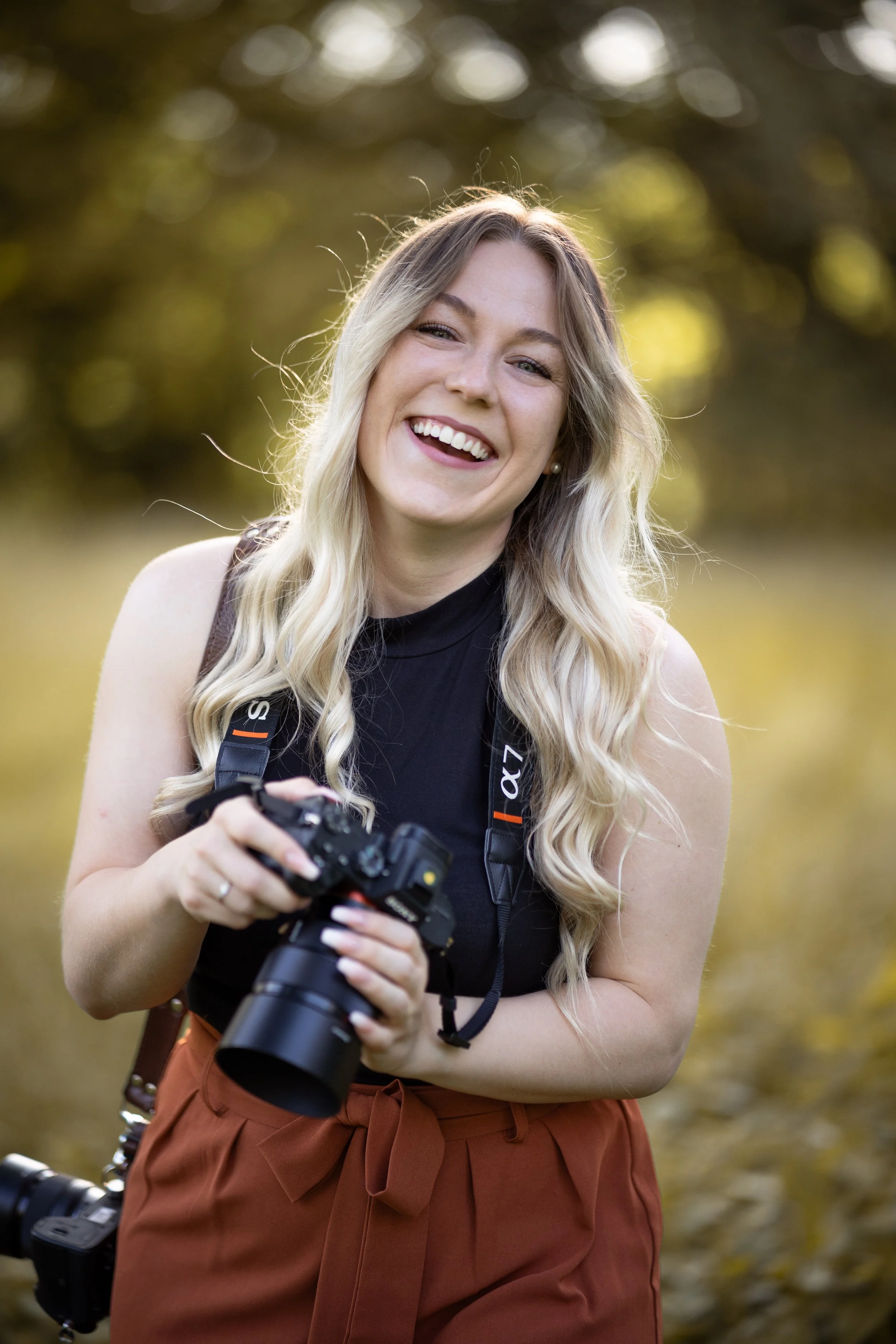 Die Fotografin, eine lachende junge Frau mit langem, blonden, gewelltem Haar hält eine Kamera vor sich. Sie steht im Freien vor einem unscharfen Hintergrund mit warmem, goldenem Sonnenlicht, während sie selbst ein Shooting aufnimmt.