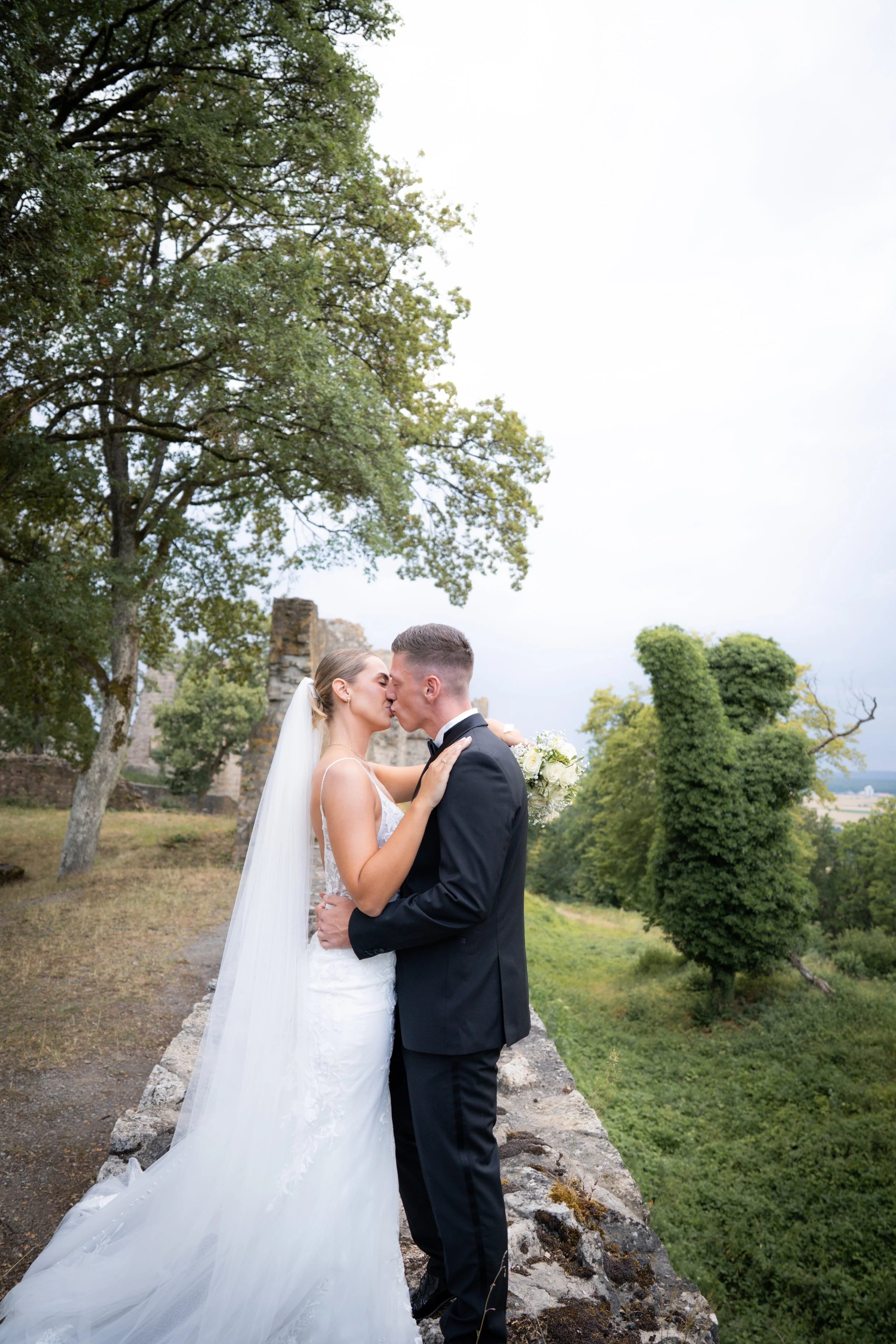 Ein Brautpaar bei einer Hochzeit im Freien, das sich küsst, umgeben von Bäumen und einer alten Burgruine im Hintergrund.
