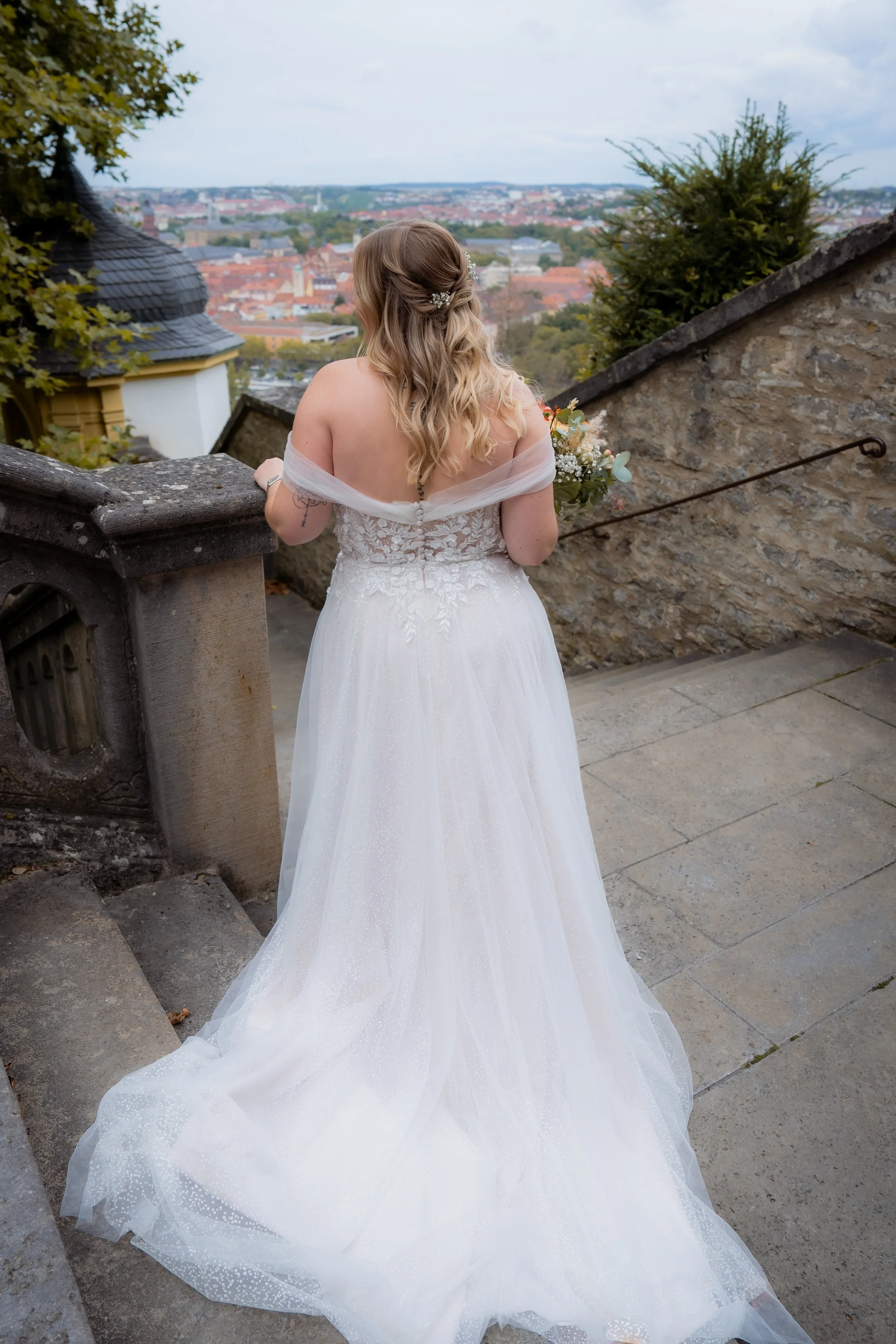Eine Braut in einem weißen Hochzeitskleid steht auf einer Treppe und schaut auf eine Stadtansicht im Hintergrund.