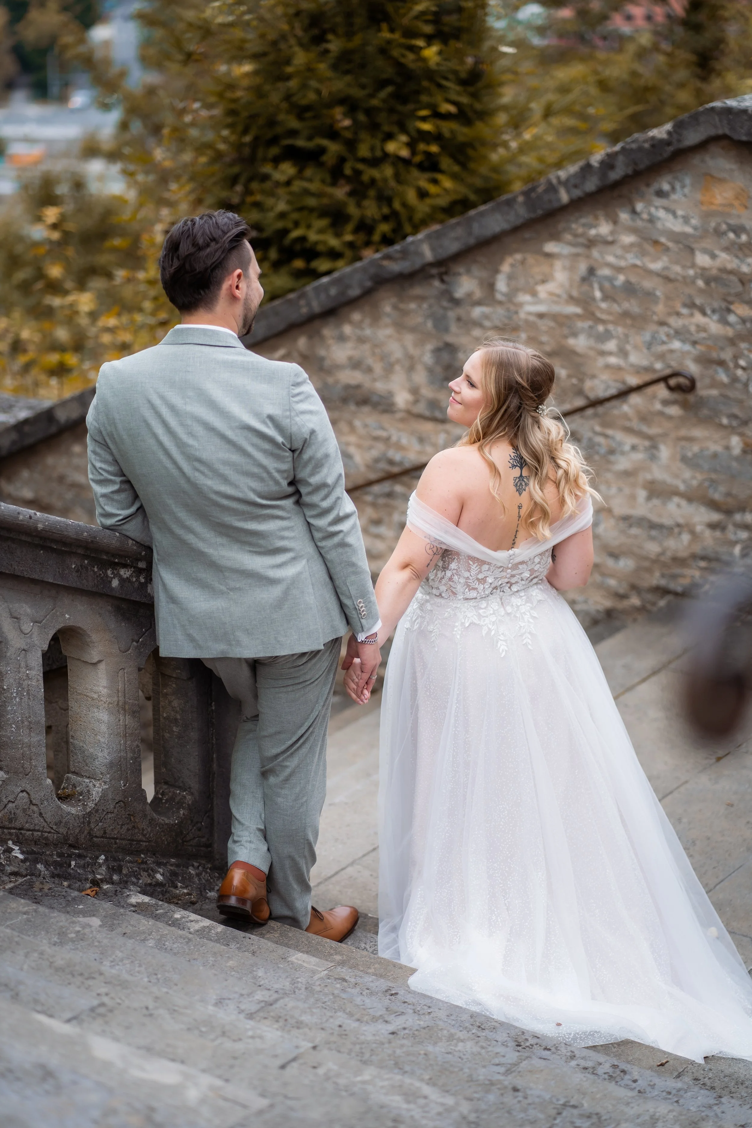 Ein Mann in einem grauen Anzug und eine Frau in einem weißen Brautkleid stehen auf einer Treppe, halten sich an den Händen und schauen sich liebevoll an, während im Hintergrund Herbstbäume und ein Steinmauer sind.