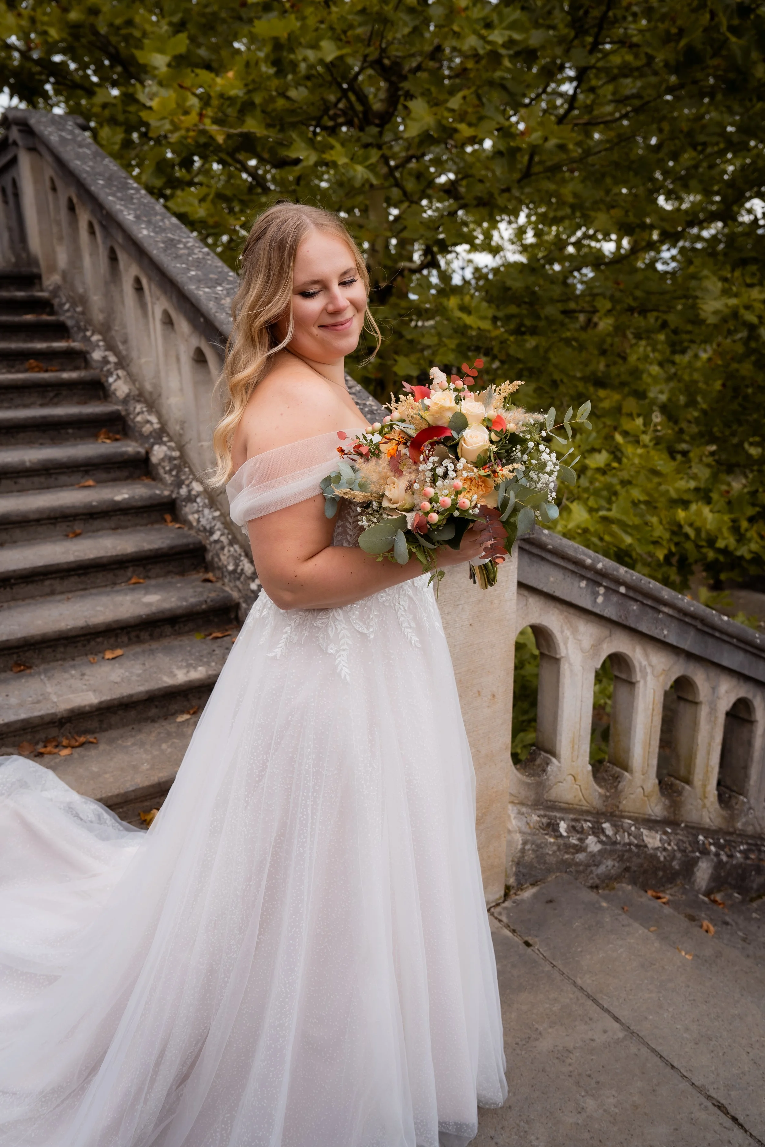 Braut in einem weißen Hochzeitskleid hält einen Blumenstrauß und steht auf einer Treppe im Freien, umgeben von grünen Bäumen.