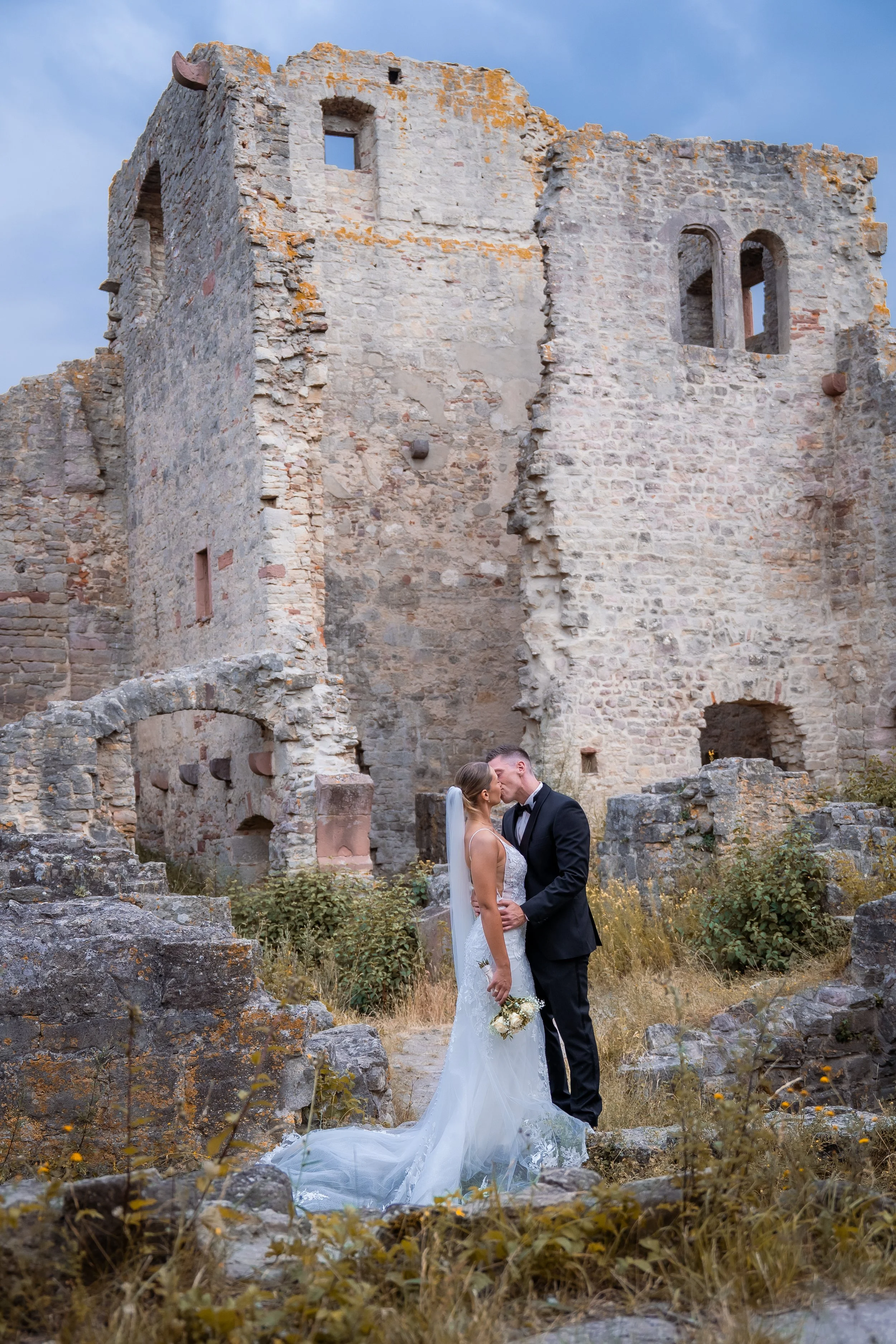 Ein heiratendes Paar in formeller Kleidung, das sich vor einer alten, zerfallenen Burg küsst, umgeben von Natur und aufgewuchertem Gelände.