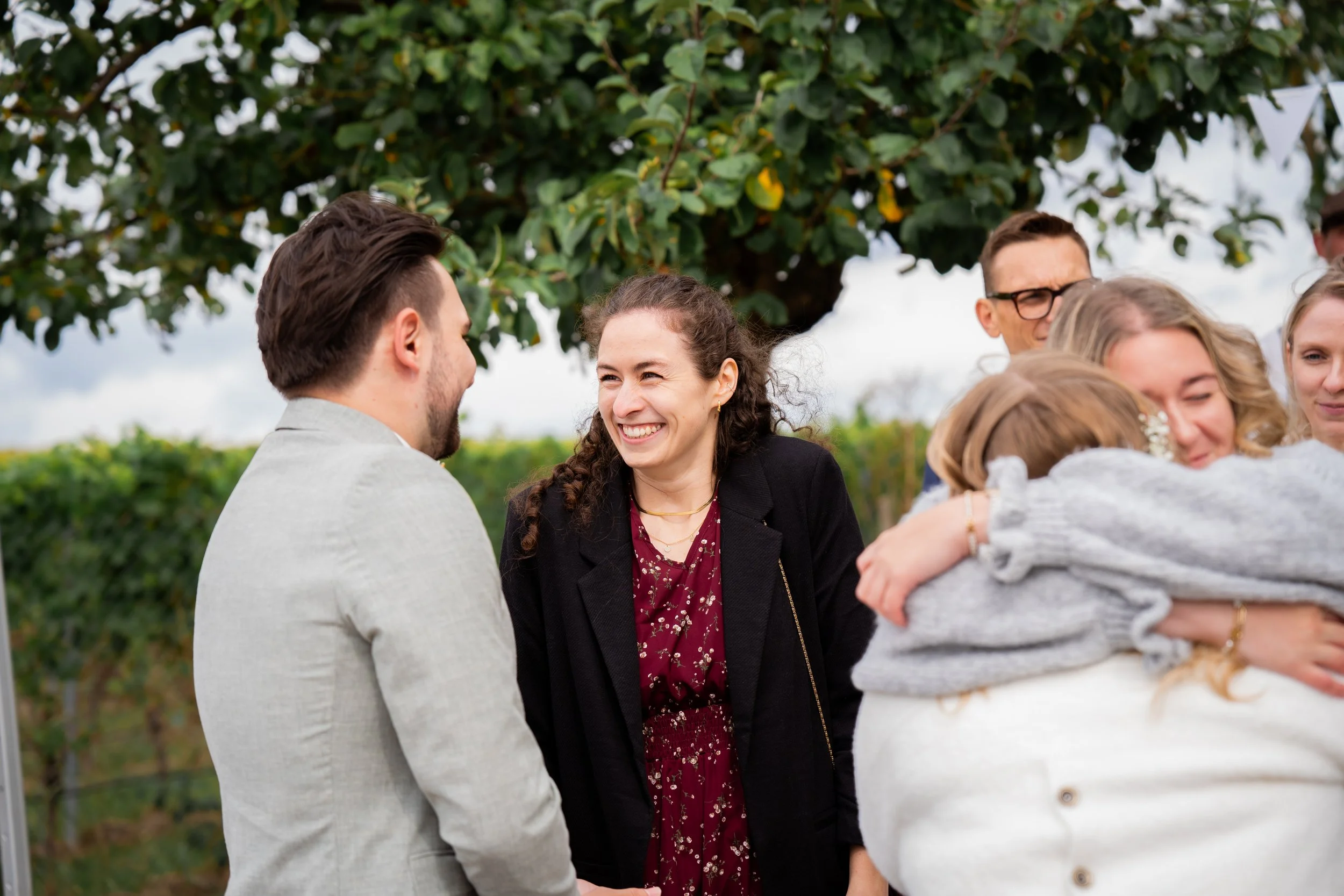 Mehrere Menschen, darunter eine Frau mit braunen, lockigen Haaren und einer schwarzen Jacke, die lächelnd in Gespräch mit einem Mann mit dunklem, kurzen Haar und grauem Blazer, umgeben von weiteren Personen in einem Garten oder Park mit großen Bäumen