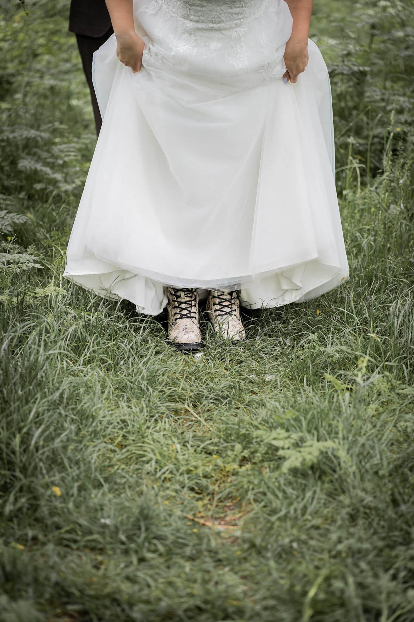 Hochzeitspaar steht im Grünen, die Braut trägt ein weißes Kleid und pinkfarbene Schuhe, die Füße der Braut sind sichtbar, während sie einen weißen Rock hochhält