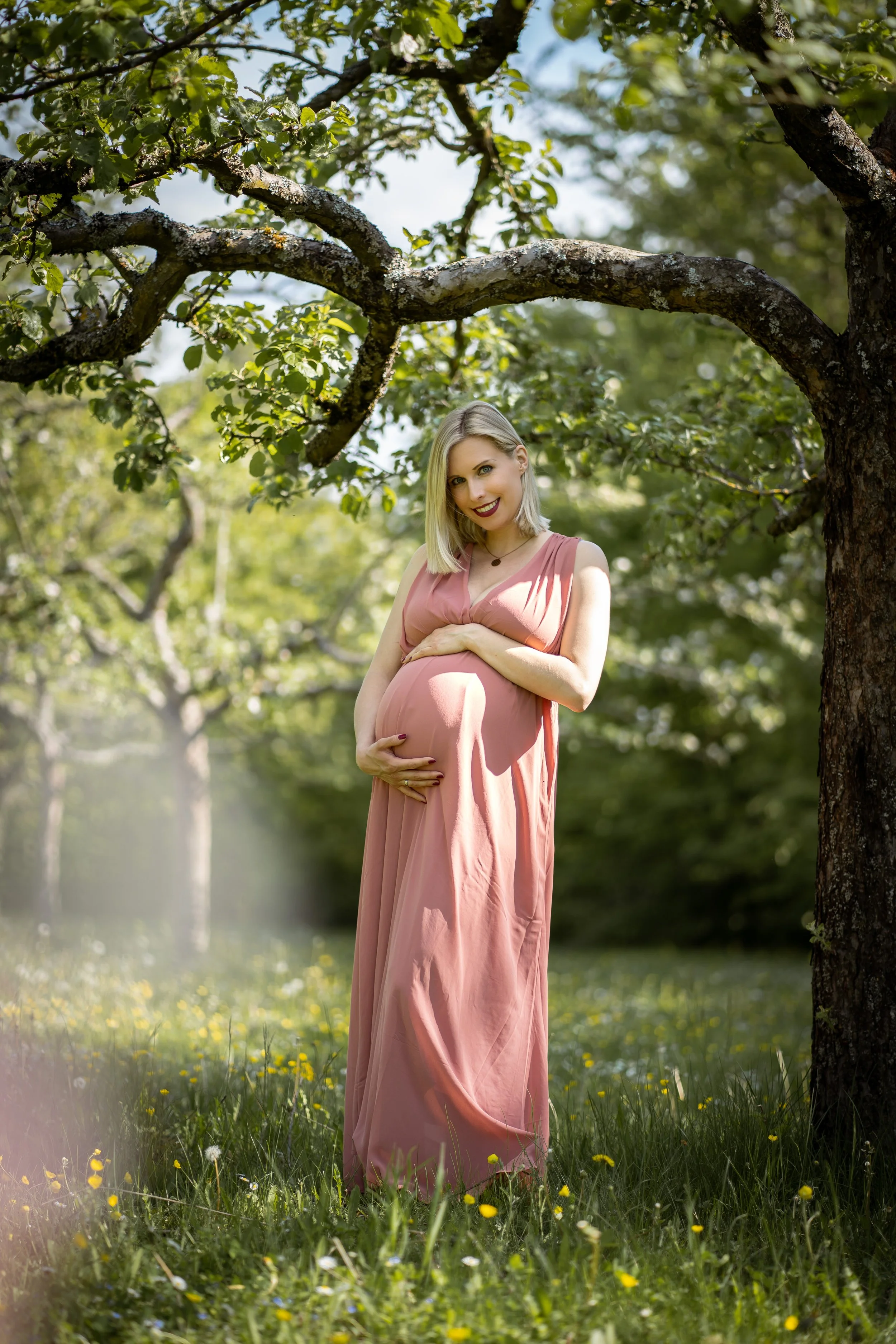 Schwangere Frau in einem langen rosa Kleid steht im Freien unter einem Baum, hält ihren Bauch und lächelt.