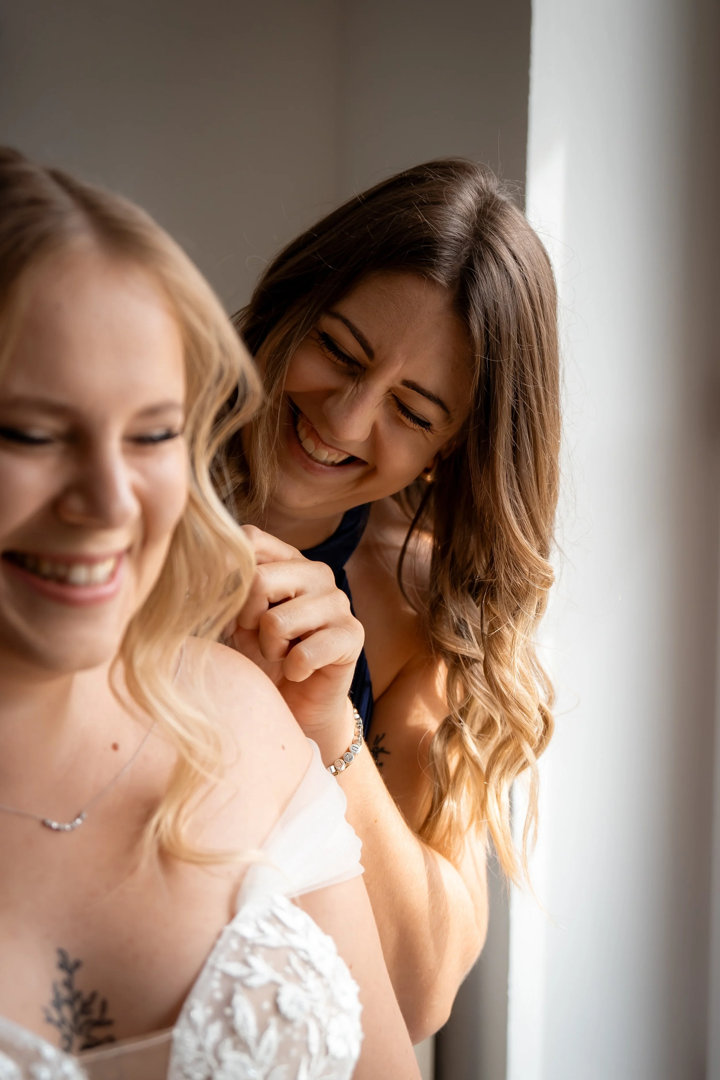 Zwei lachende Frauen, eine blonde Frau in weißem Kleid und eine braunhaarige Frau, die ihr eine Spritze gibt, vermutlich beim Impfen.