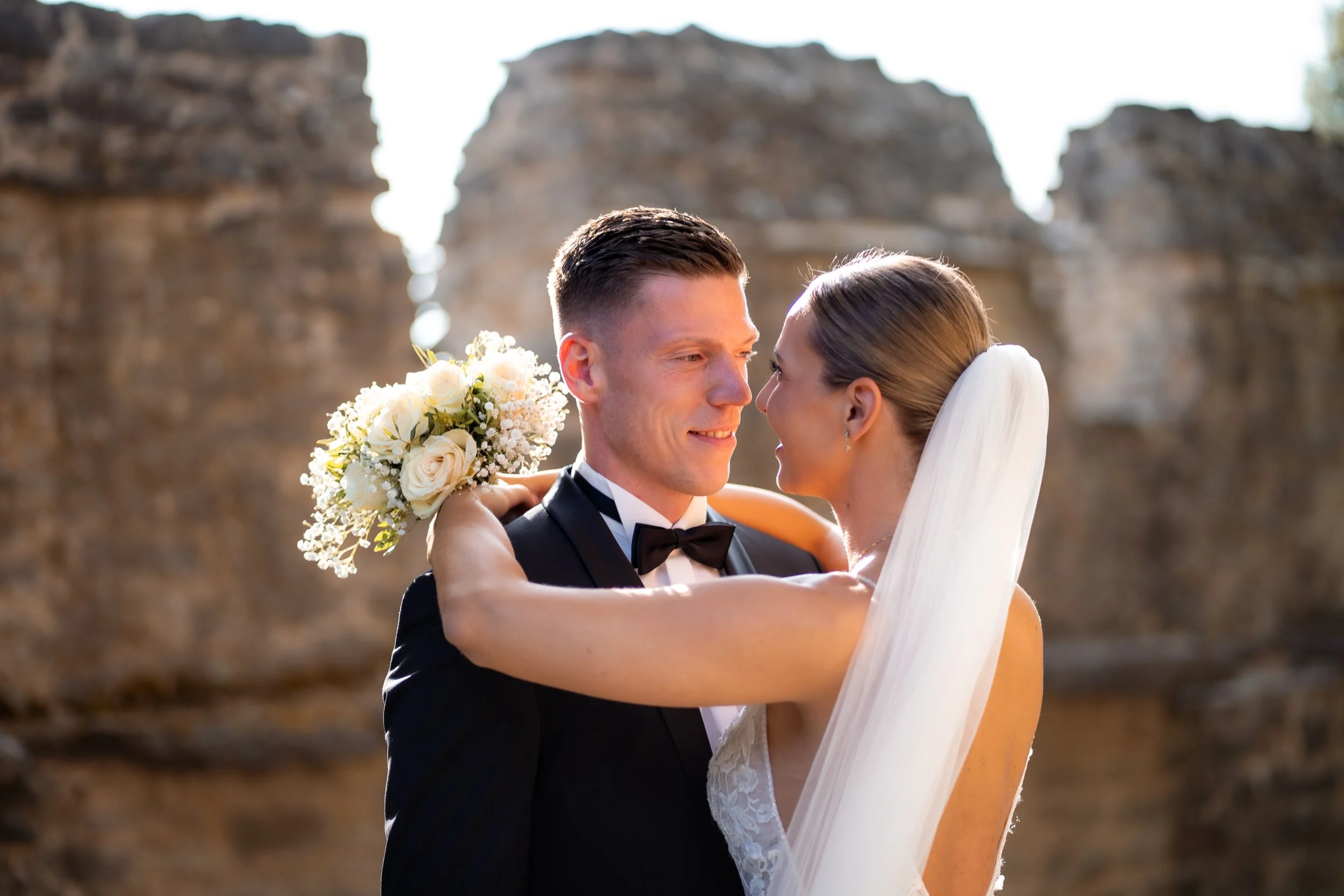 Ein glückliches Brautpaar bei einer Hochzeit im Freien, der Bräutigam trägt einen schwarzen Anzug mit Fliege, die Braut trägt ein weißes Kleid mit Schleier, sie umarmt sich liebevoll, im Hintergrund sind alte Felsen und Himmel sichtbar.