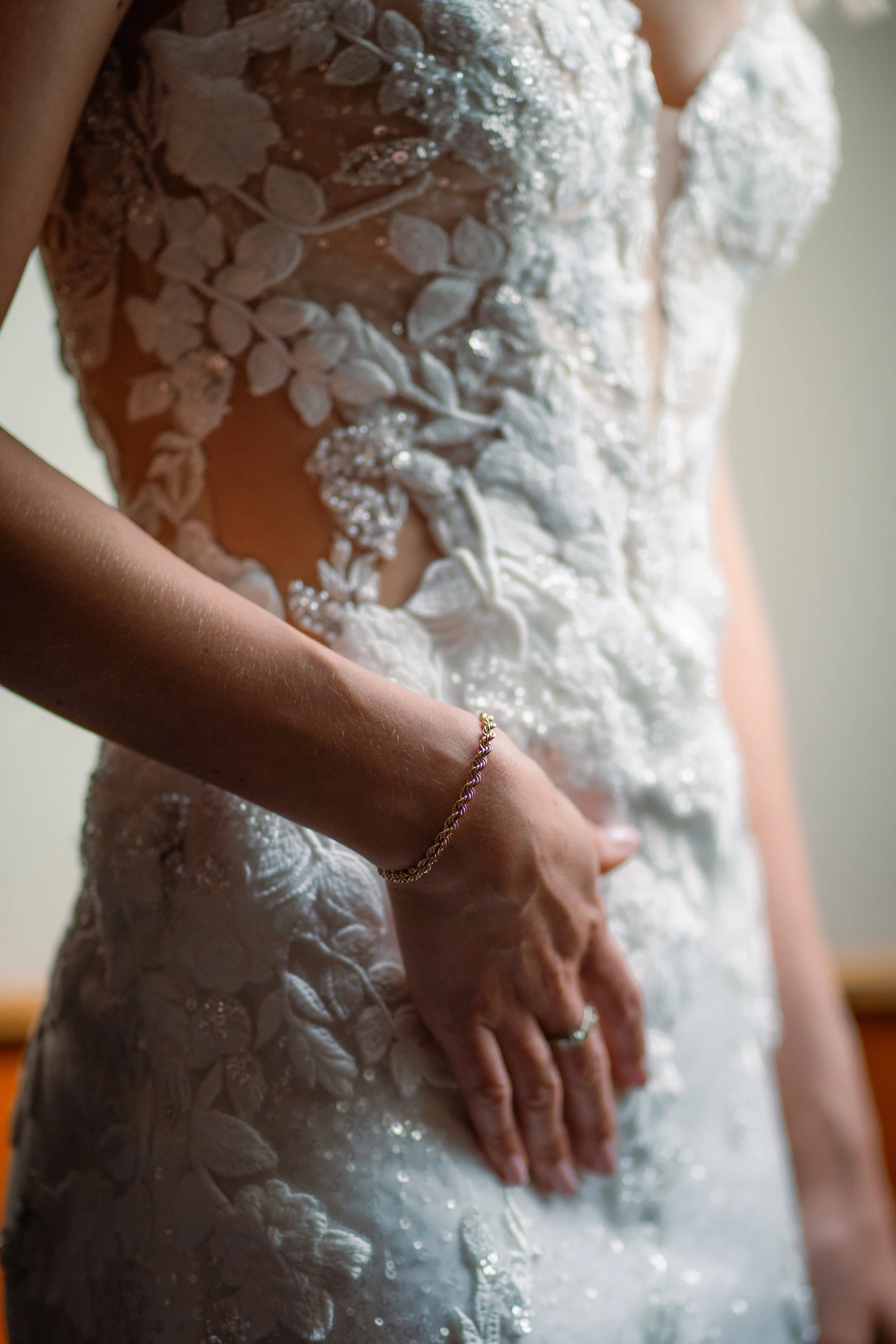 Nahaufnahme eines Brautkleides mit floraler Applikation und Edelsteinen, die am Oberkörper einer Braut sichtbar sind, während sie ihre Hand mit einem Ring hält.