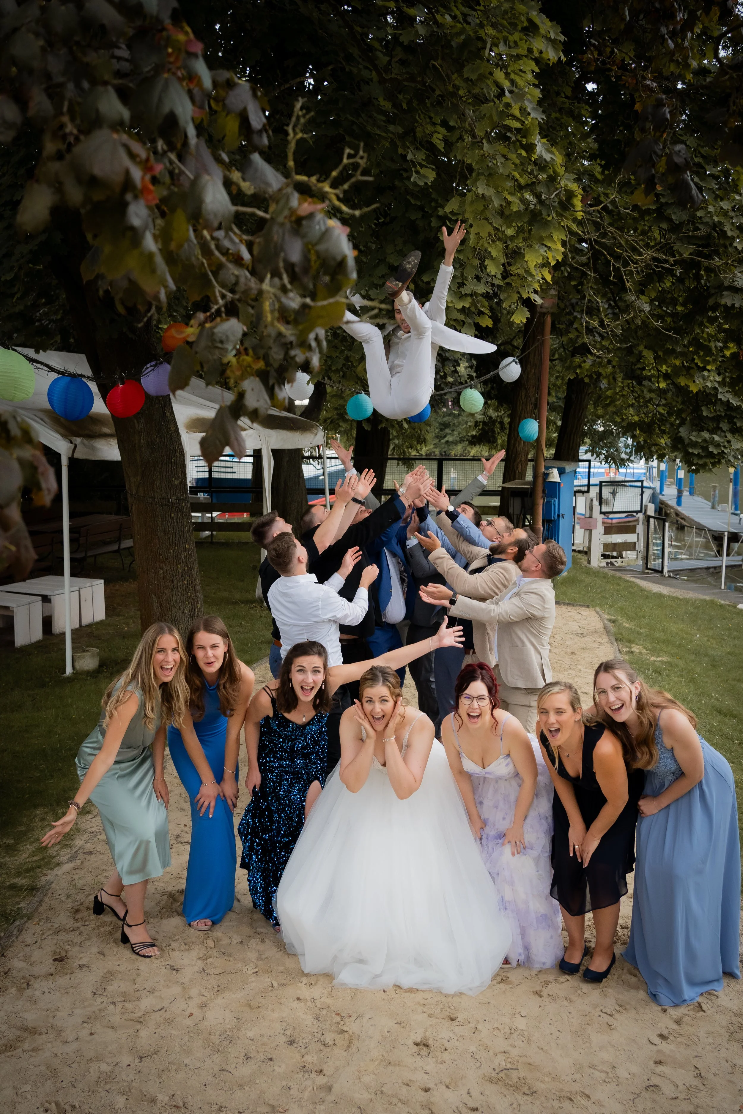 Feierliche Gruppe von Menschen bei einer Hochzeit, einige werfen die Braut in die Luft, im Hintergrund sind Bäume und Lichterketten sichtbar.