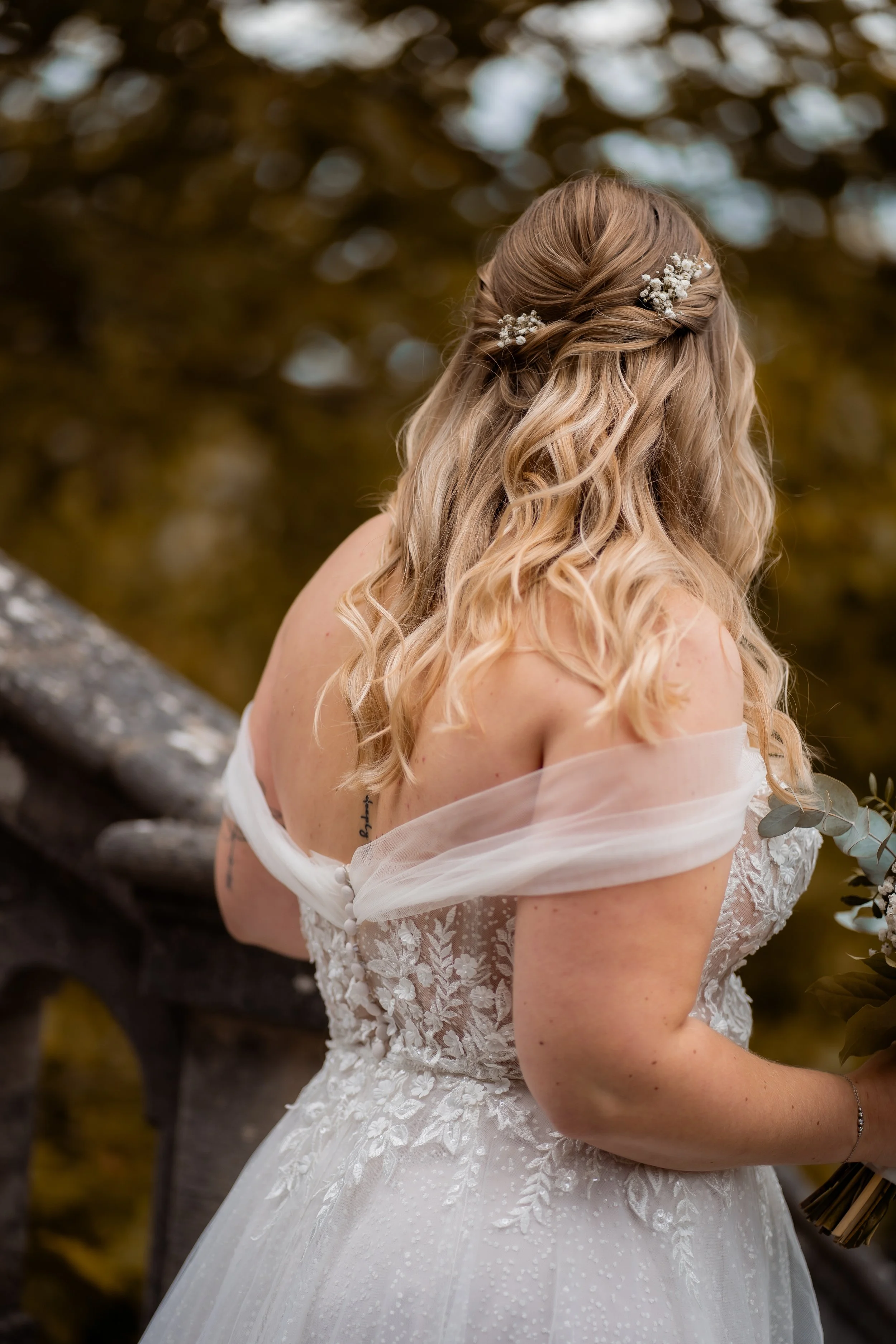 Braut trägt ein weißes Hochzeitskleid mit floraler Stickerei, ihre blonden, lockigen Haare sind mit kleinen weißen Blumen geschmückt, sie steht an einem Baum mit herbstlichen Blättern im Hintergrund.