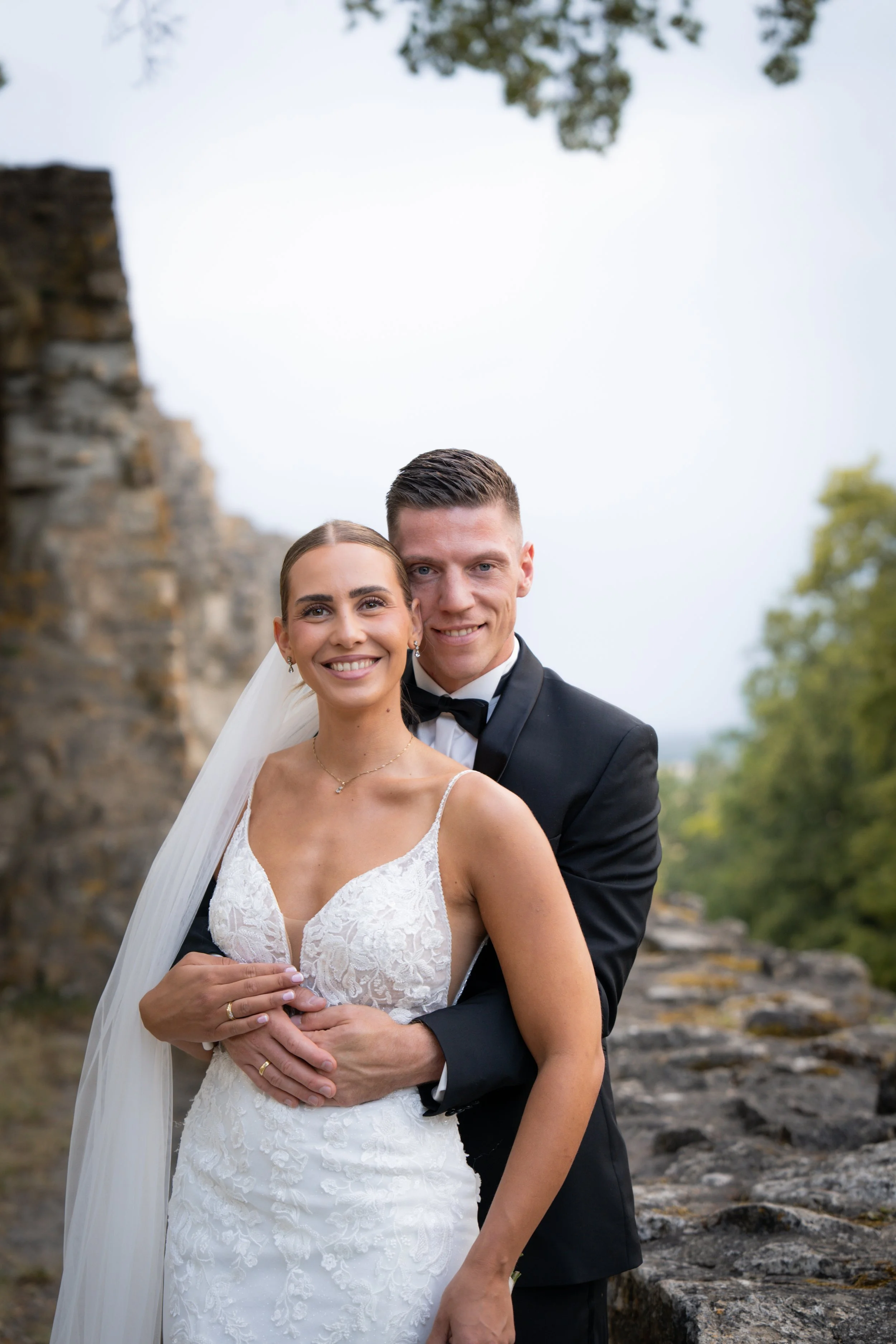 Ein glückliches Brautpaar steht im Freien, die Braut trägt ein weißes, spitzenbesetztes Hochzeitskleid und die Hochzeitskette, der Bräutigam trägt einen schwarzen Anzug mit Fliege. Sie stehen vor einer alten Steinmauer und umgeben von grünen Bäumen.