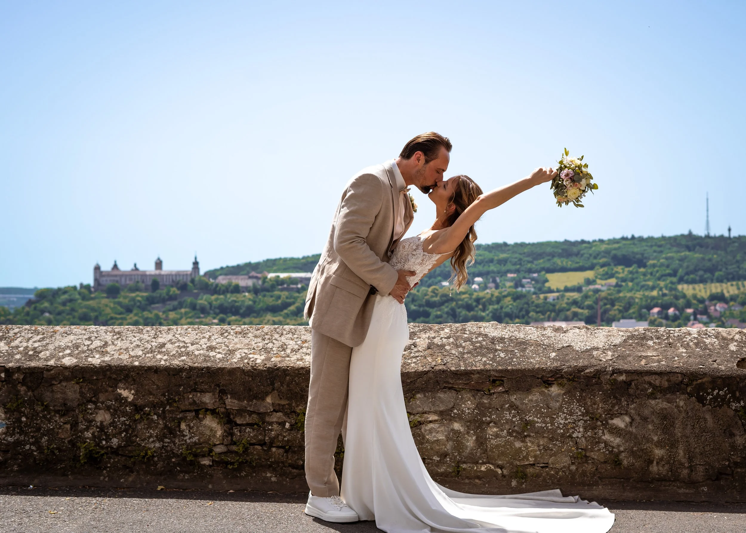Ein Paar bei einer Hochzeit, der Bräutigam küsst die Braut, die die Arme in die Luft hebt und einen Blumenstrauß hält, vor einer alten Mauer mit Stadt- und Hügelhintergrund bei schönem Wetter.