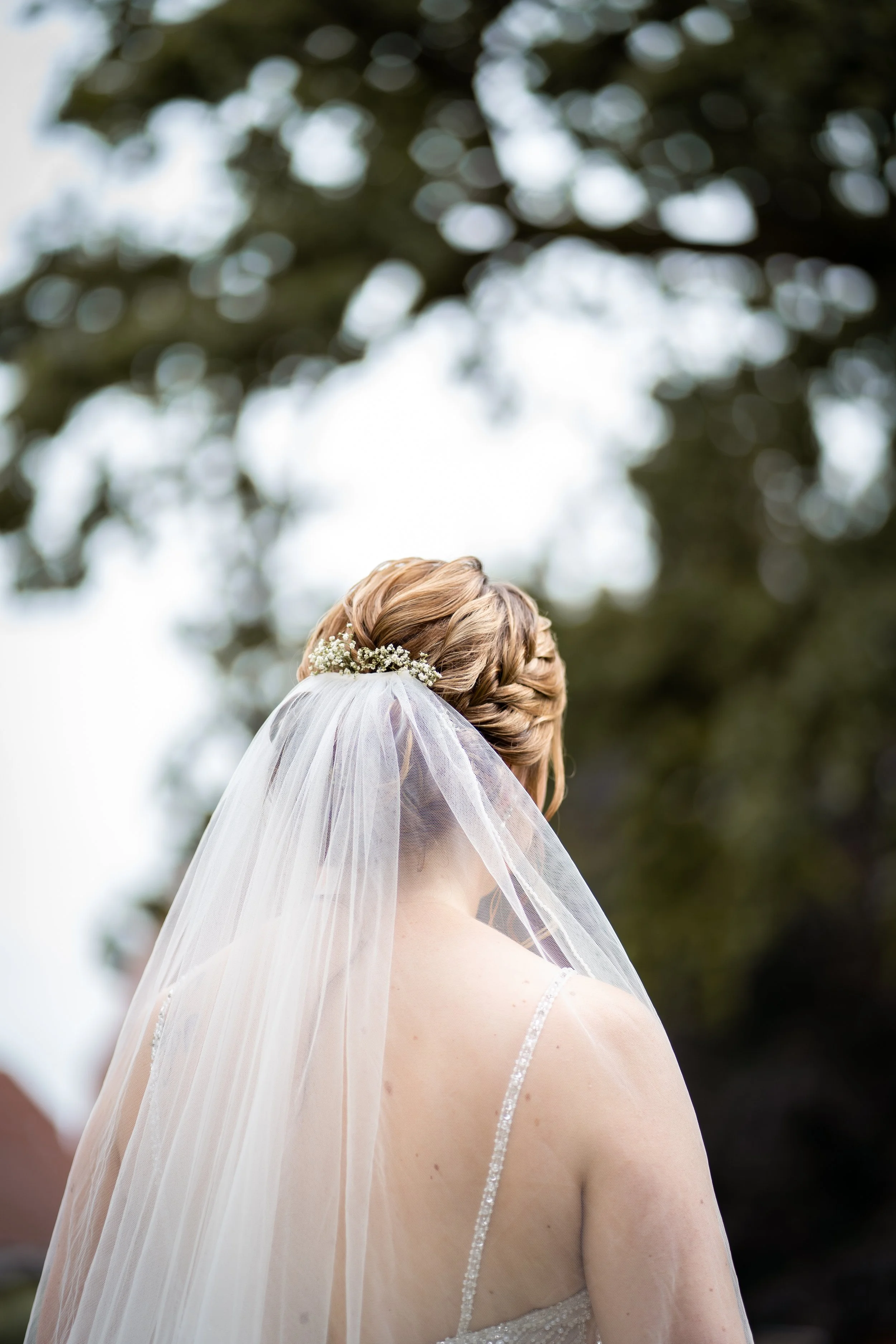 Nahaufnahme eines Brautkleids mit Schleier, Hochzeitsfrisur und Blumen im Haar, im Freien mit Bäumen im Hintergrund.