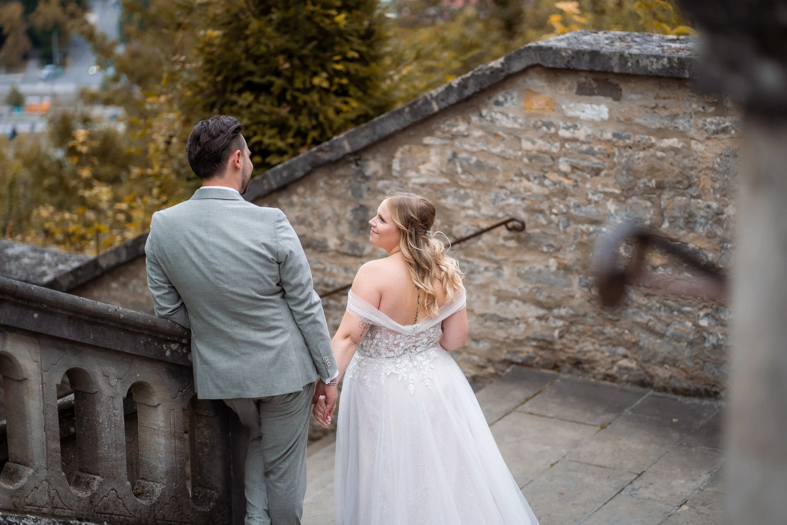 Ein Brautpaar im Hochzeitsoutfit hält Händchen auf einer Steinbrücke im Park, umgeben von Herbstbäumen.